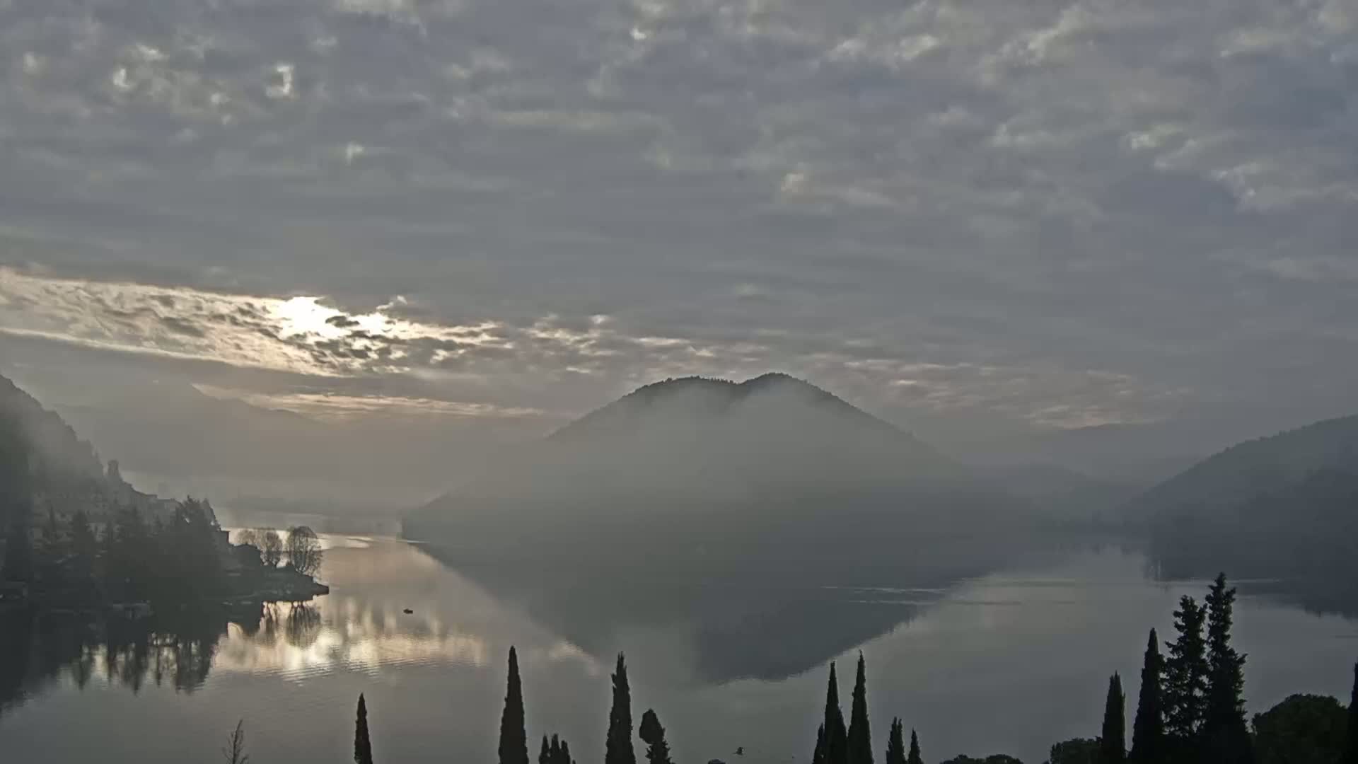 A calm lake is shrouded in mist at sunrise, with hills and trees lining the shore under a cloudy sky.