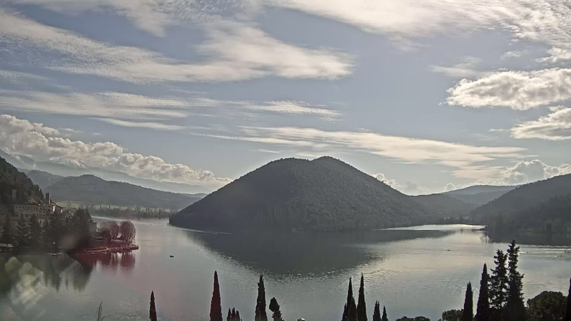 A calm lake reflects a partly cloudy sky and is surrounded by hills and a forested island, with a small village visible on the shoreline.