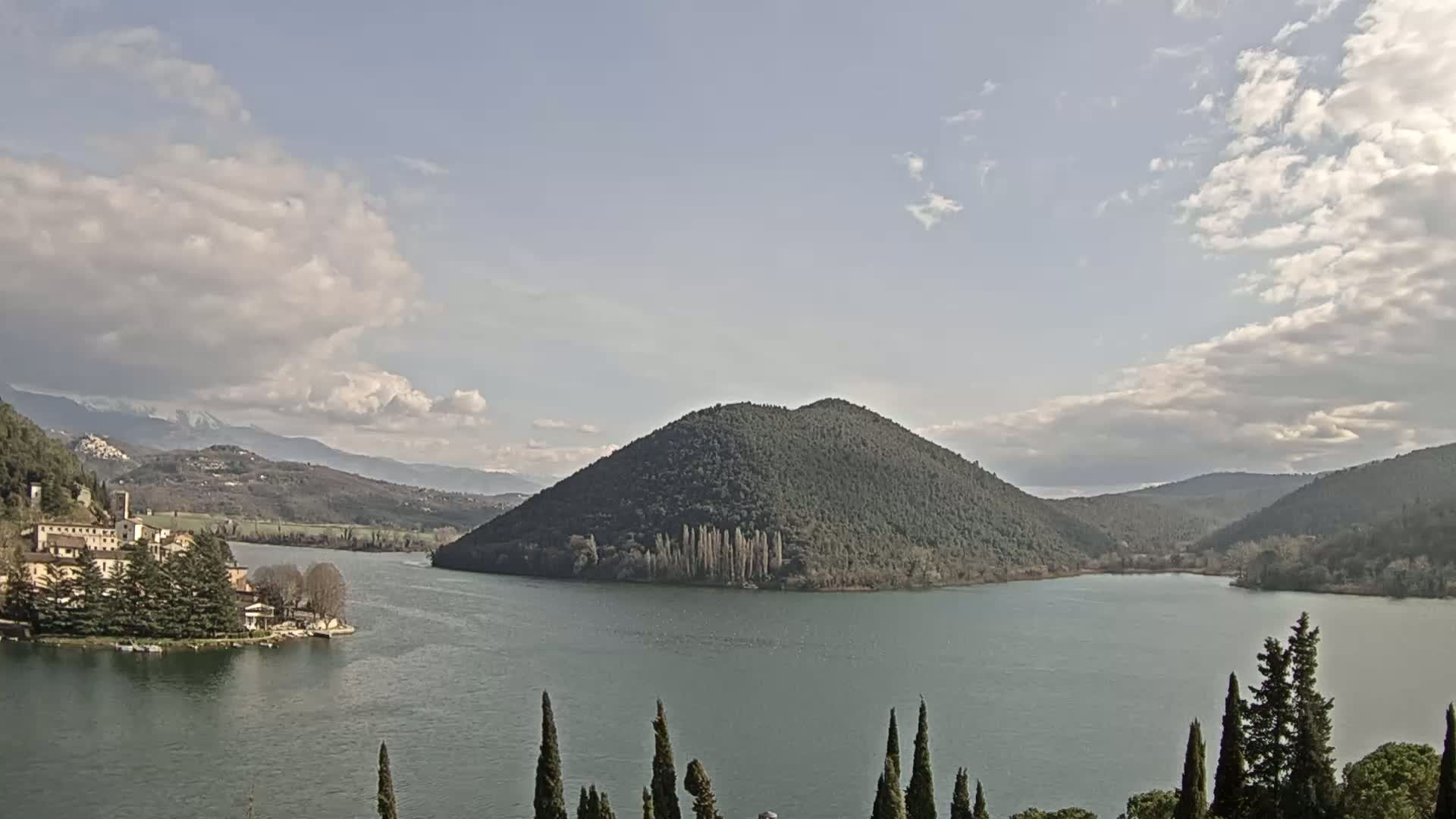 A partly cloudy day reveals a tranquil lake surrounded by green hills and a small village nestled on its shore.