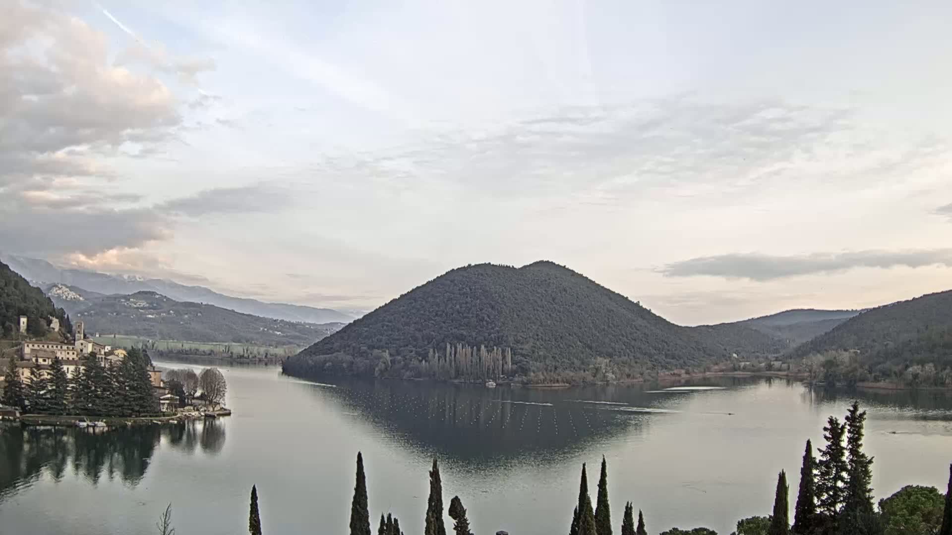 A lake is nestled between tree-covered hills under a cloudy sky, with a village and snow-capped mountains visible in the distance.