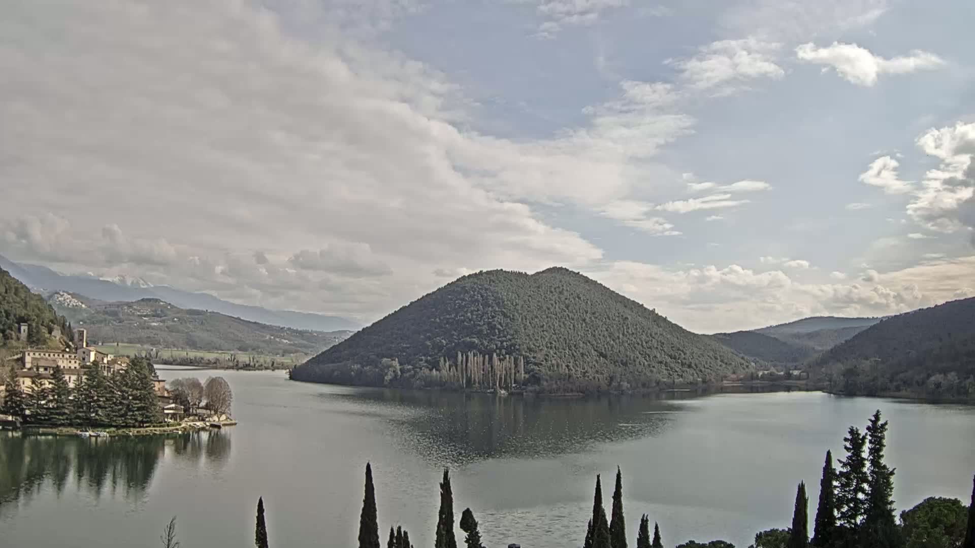 A lake is surrounded by hills and mountains under a partly cloudy sky, with a village visible on the lake's shore.