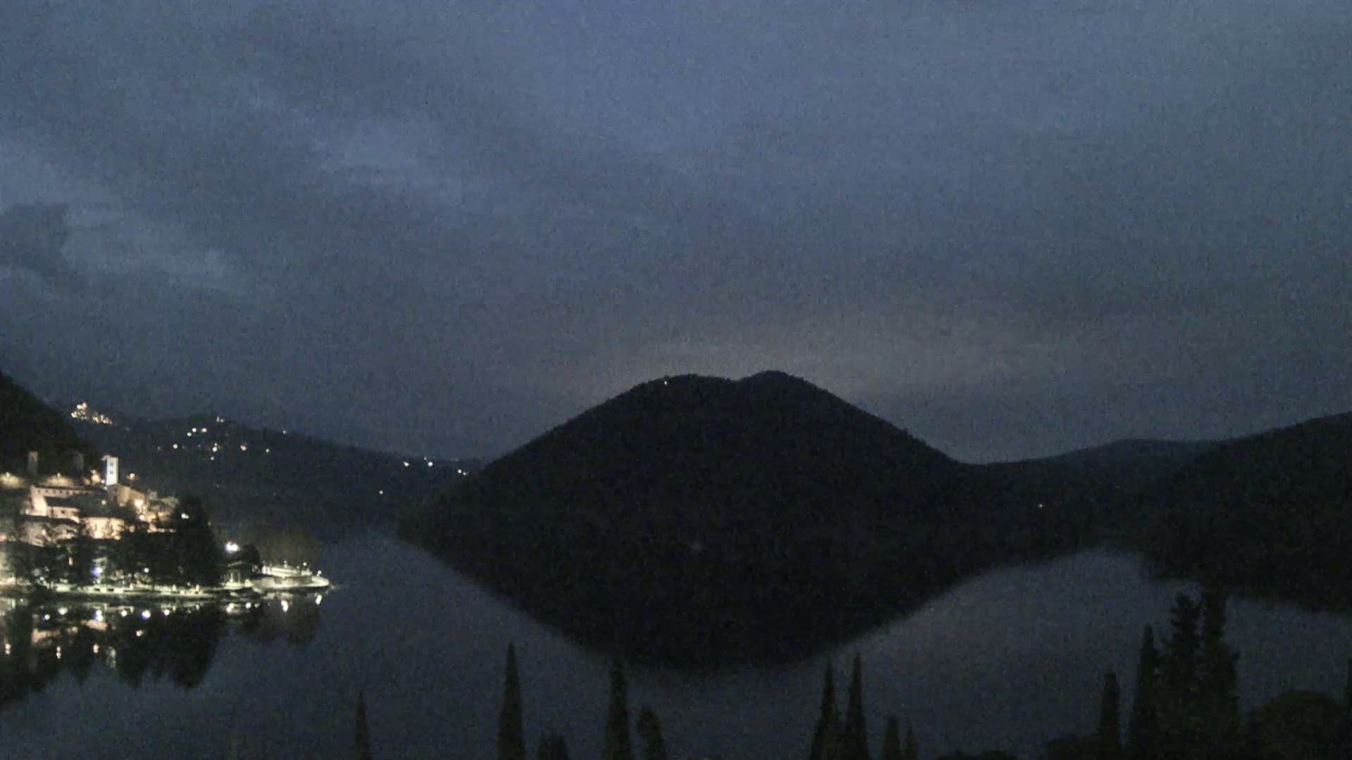 A dark, calm lake reflects a hill and a small, lit town under a dusky sky.