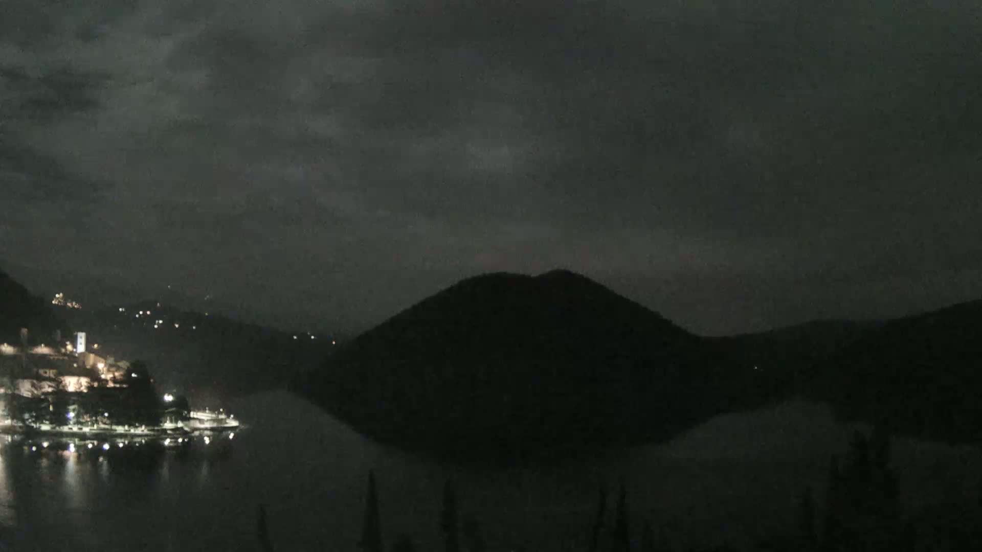 A nighttime view of a dark, hilly landscape with a town's lights reflecting on a calm body of water under a cloudy sky.