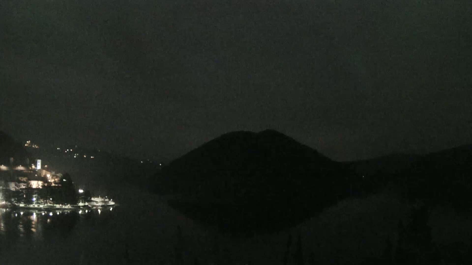 A dark, nighttime scene shows a body of water reflecting the lights of a small town nestled between hills under a cloudy sky.