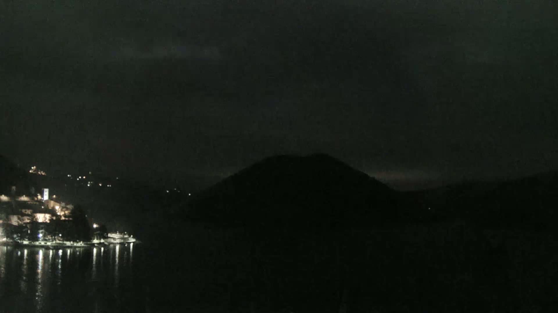 A dark nighttime scene shows a body of water reflecting scattered lights from a town nestled on a hillside, with a large, dark hill rising from the water in the background under a cloudy sky.