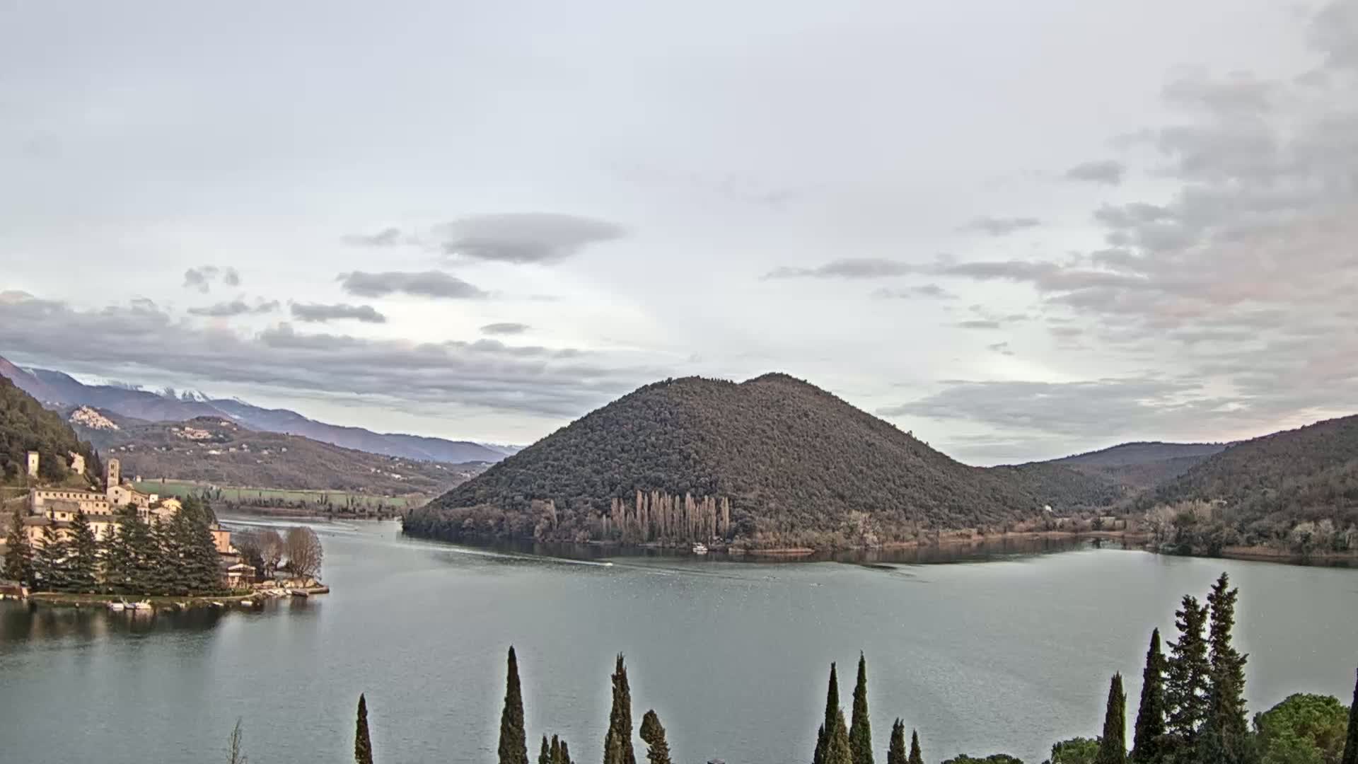 A lake surrounded by hills and mountains under a cloudy sky, with a small village and island visible in the distance.