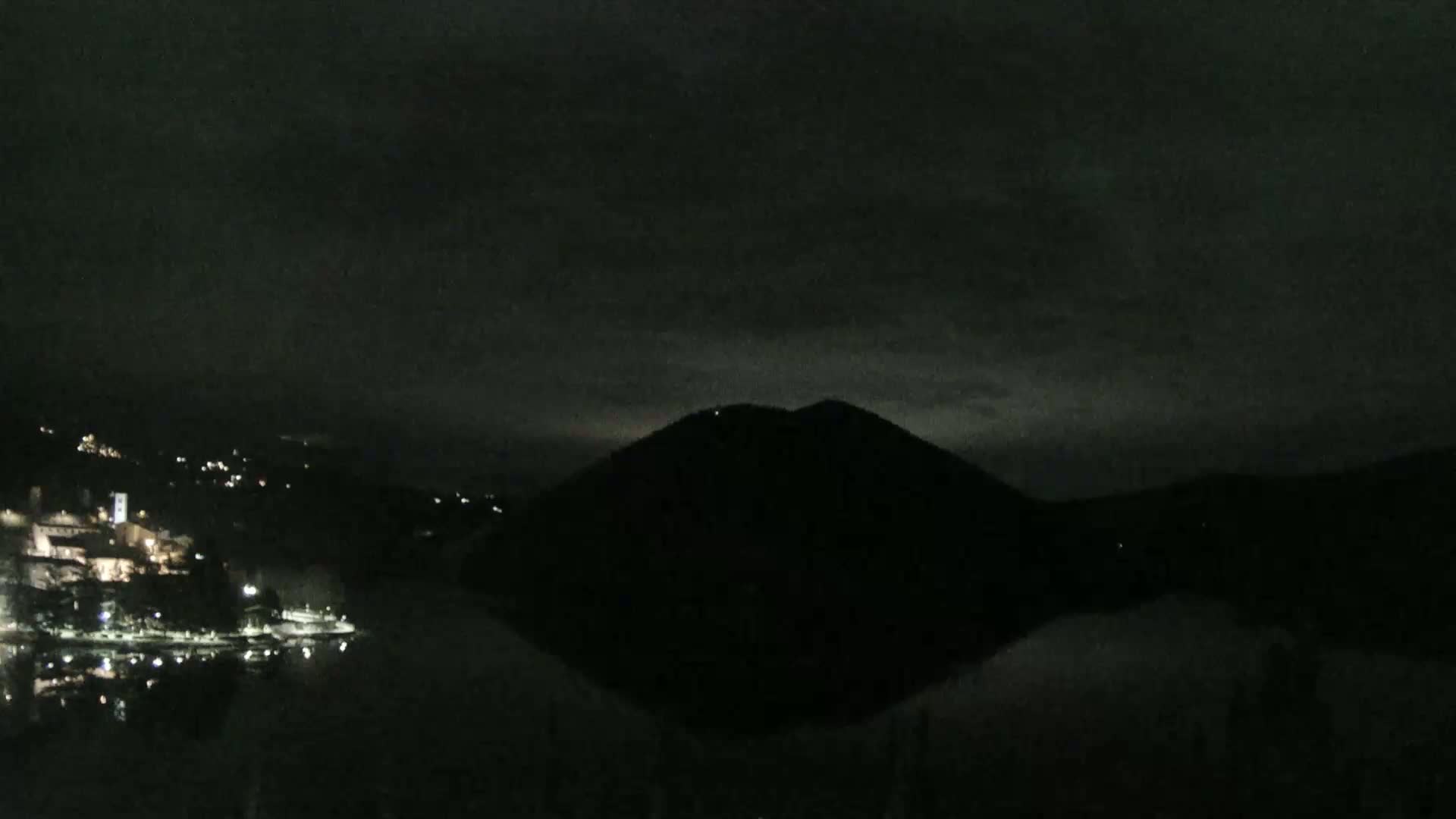 A dark, nighttime scene shows a body of water reflecting a silhouetted hill under a cloudy sky, with faintly lit buildings visible on the opposite shore.