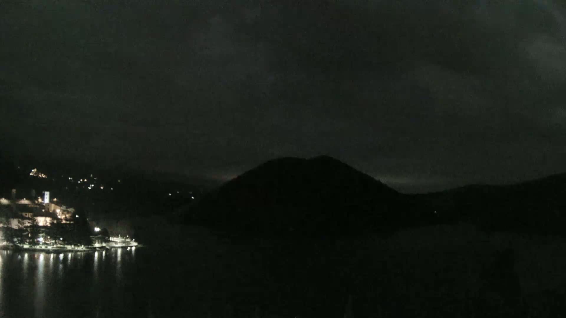 A nighttime view of a dark, mountainous landscape with a town's lights visible along a dark body of water under a cloudy sky.