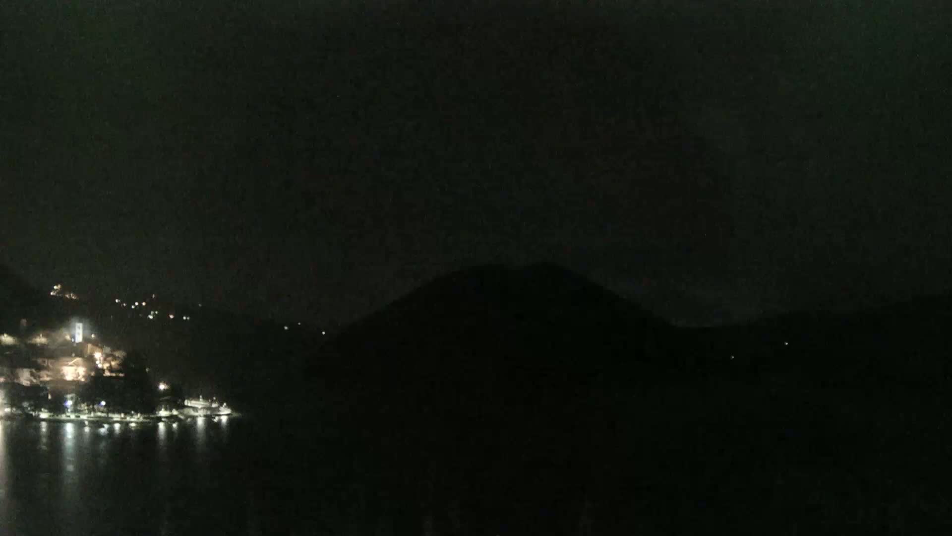 A dark nighttime scene shows a body of water reflecting scattered lights from a town nestled at the base of a large, silhouetted hill.