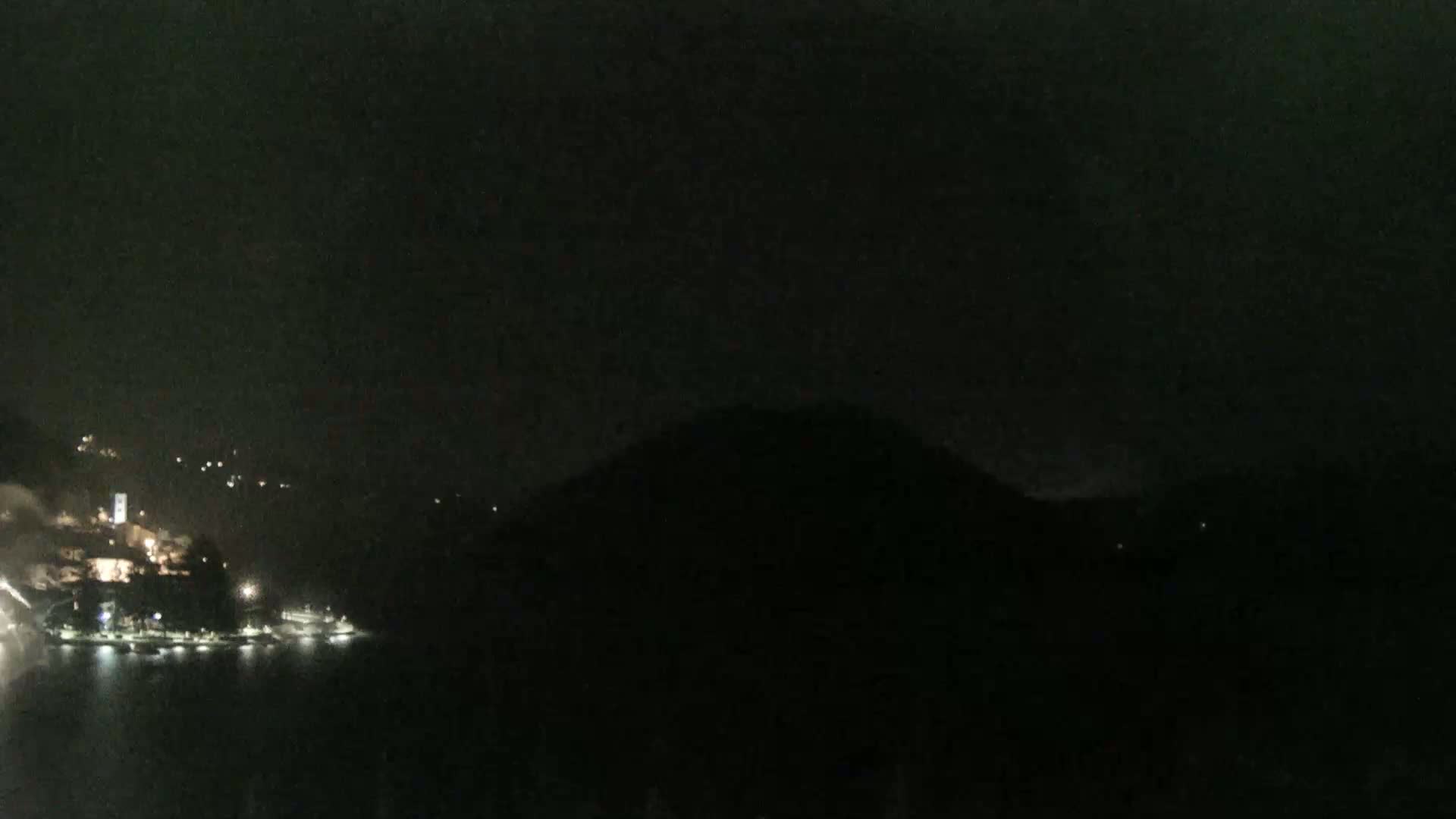 A dark nighttime scene shows a body of water with a few lights visible on the far shore, a hill silhouetted against the dark sky, and a few faint lights scattered across the hills in the background.