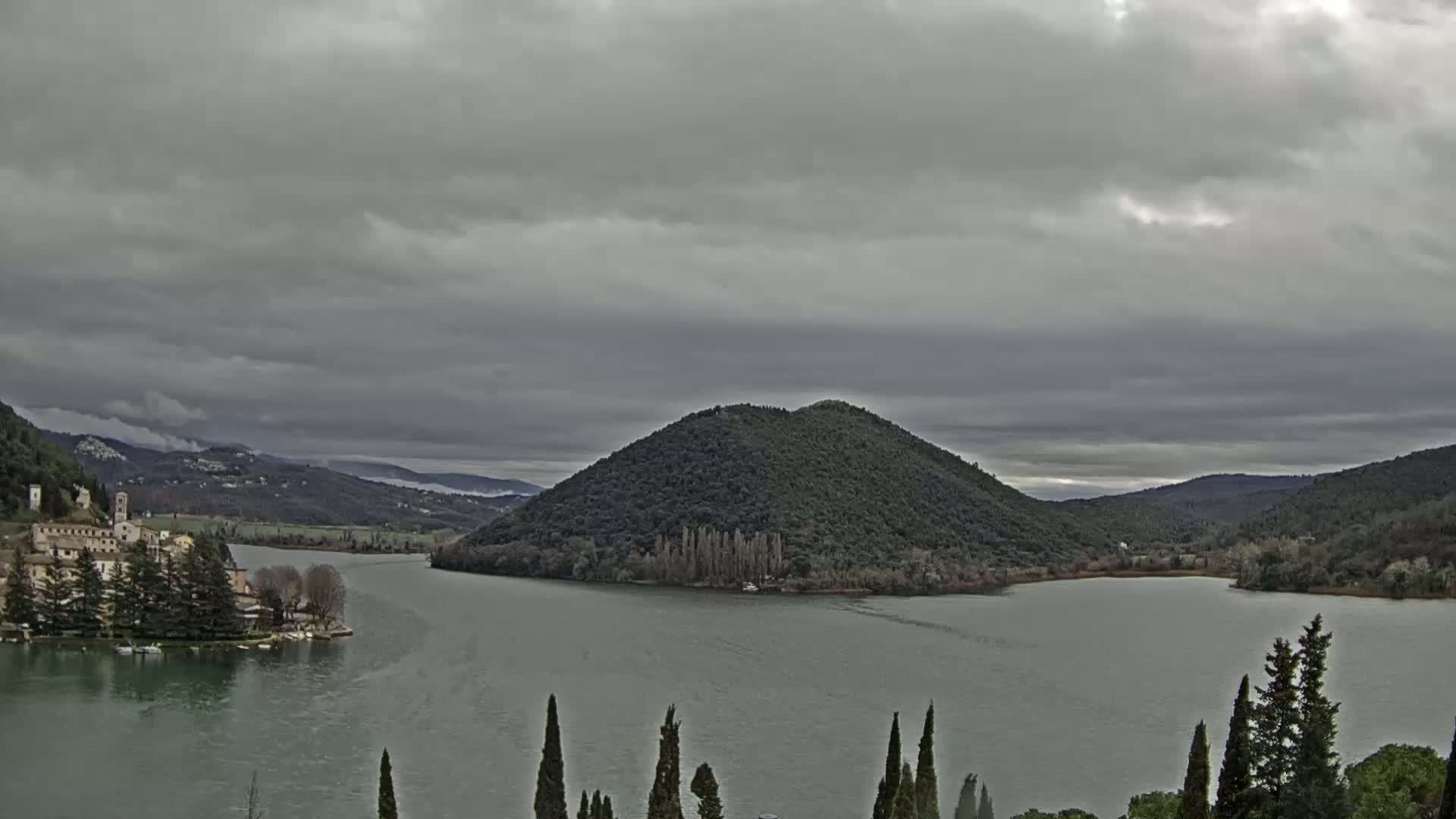 A lake, under a cloudy sky, is surrounded by hills and a small town on one shore, with a large, tree-covered hill in the middle of the lake.