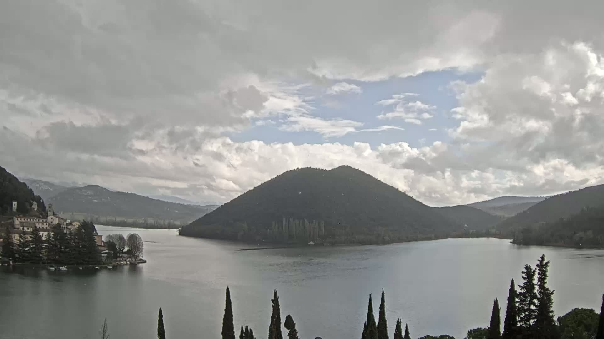 A lake is seen under a mostly cloudy sky, with a village and a large hill in the background.