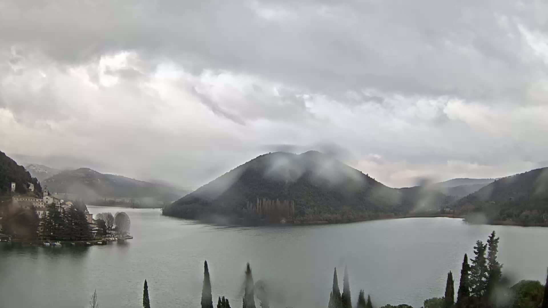 A lake surrounded by rain-covered hills and a small village is seen under a cloudy, rainy sky.