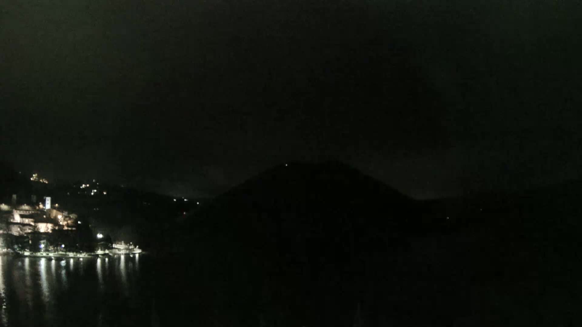 A nighttime view shows a dark hill silhouetted against a mostly clear, dark sky, with a small, lit town visible on the waterfront across a dark body of water.