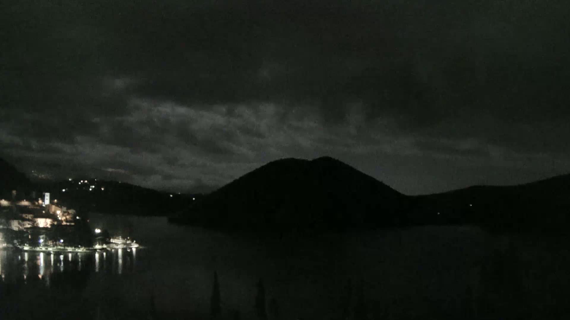 A dark, cloudy night scene shows a body of water reflecting the lights of a town on the opposite shore, with a silhouetted hill rising from the water in the midground.