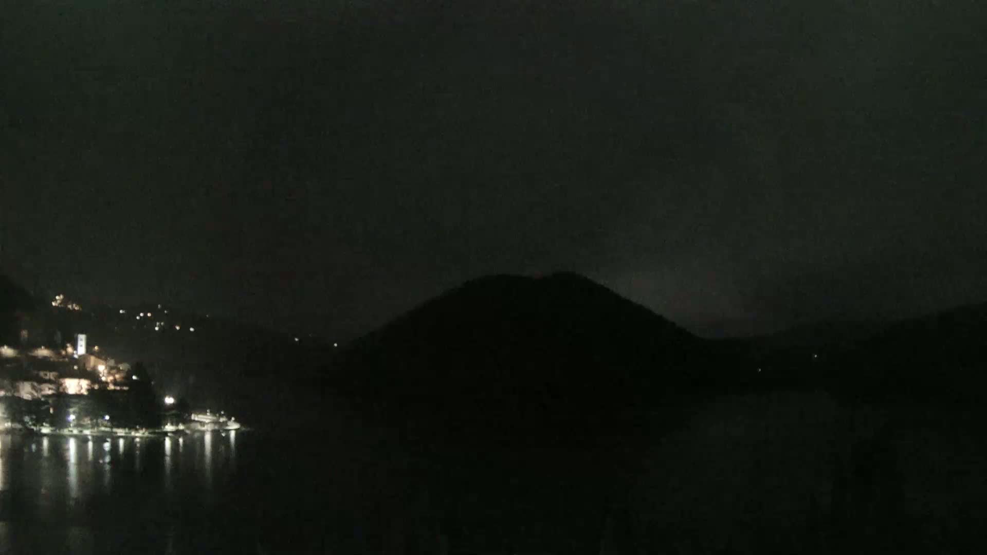 A dark, nighttime scene shows a body of water reflecting the lights of a town nestled on a hillside opposite a large, silhouetted hill.