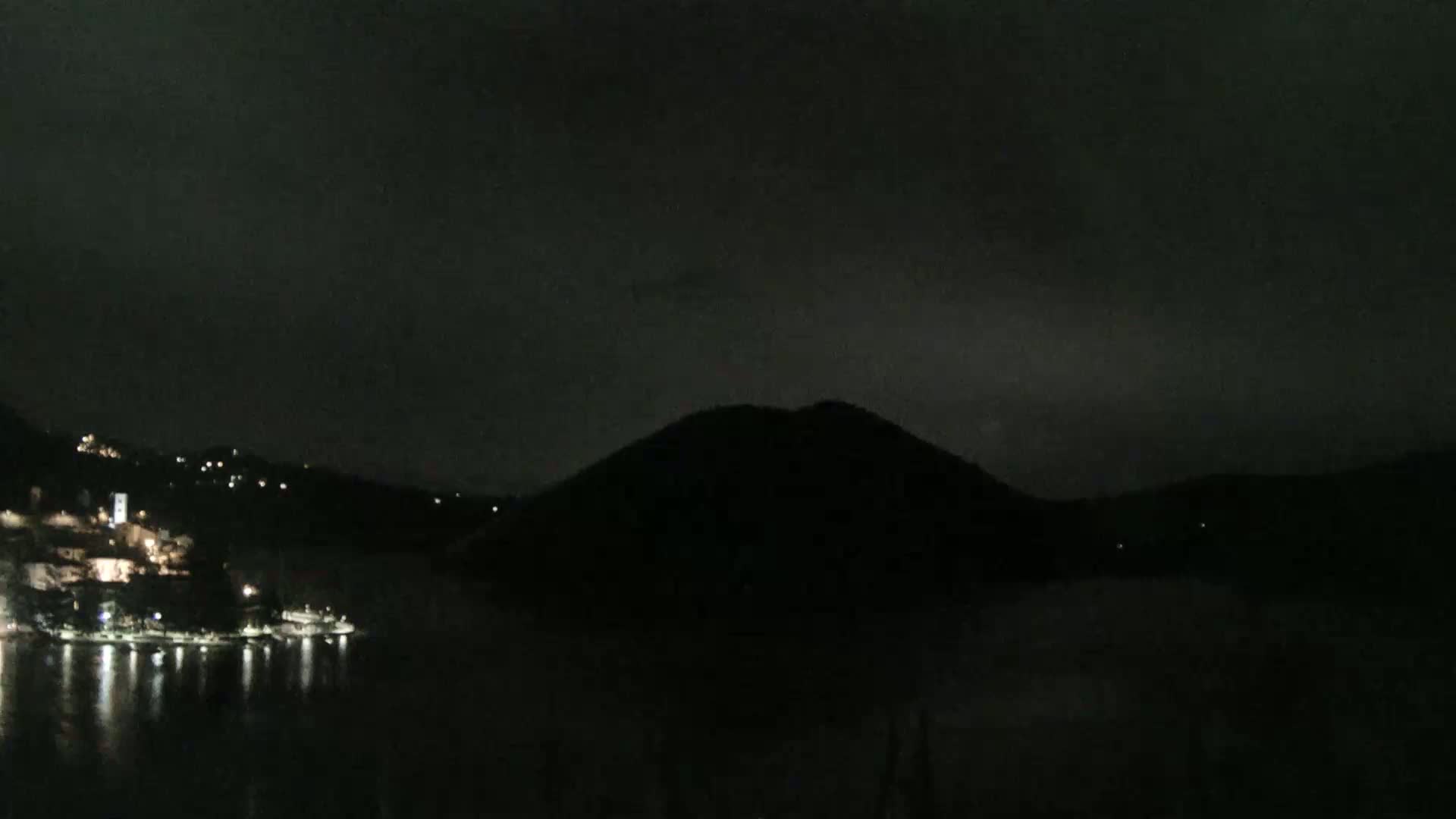 A dark, nighttime scene shows a body of water reflecting the lights of a small town on one side, with a large, silhouetted hill on the other side under a cloudy sky.