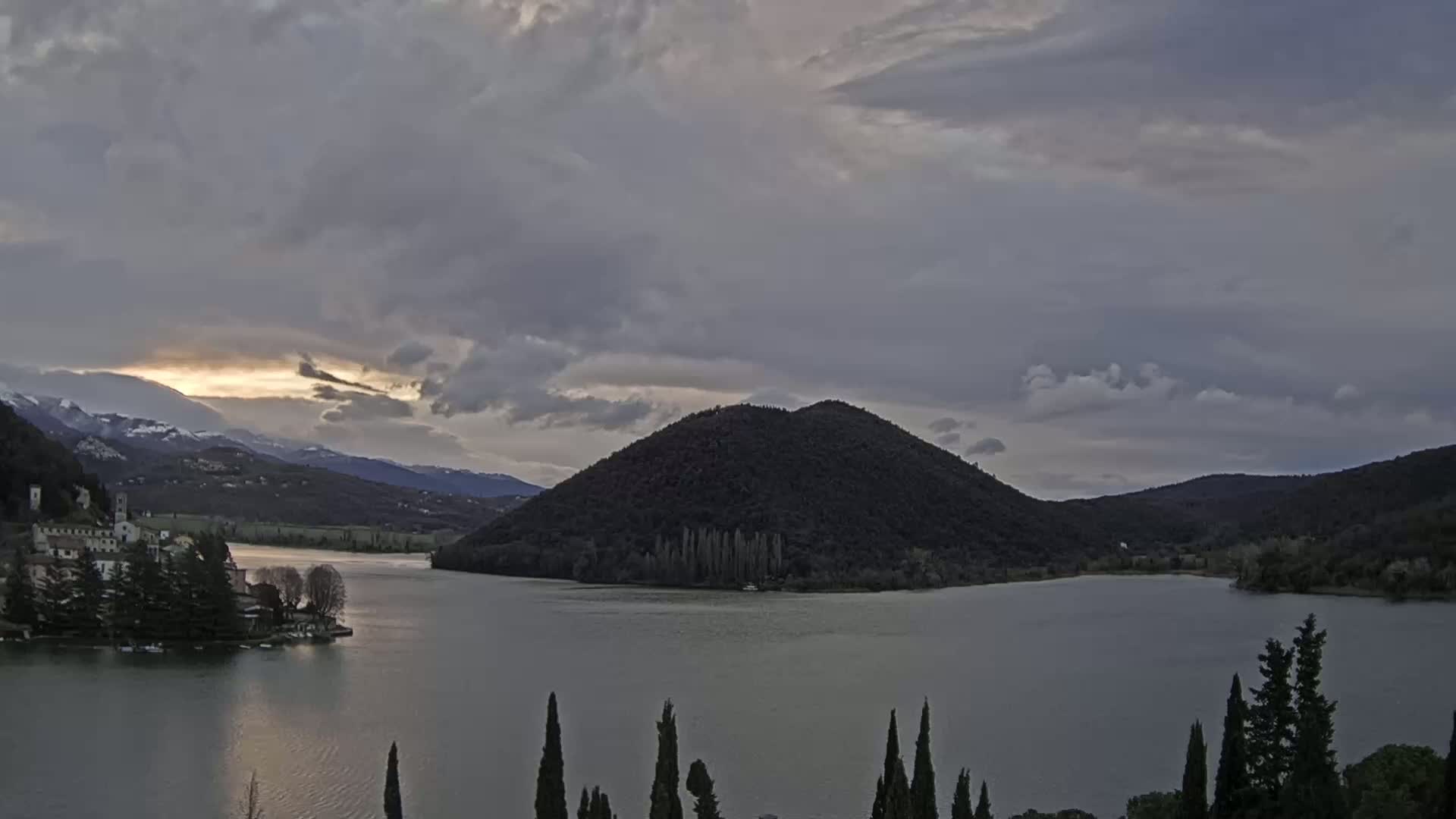 A lake surrounded by hills and mountains under a cloudy sky, with a small village on a peninsula in the foreground.
