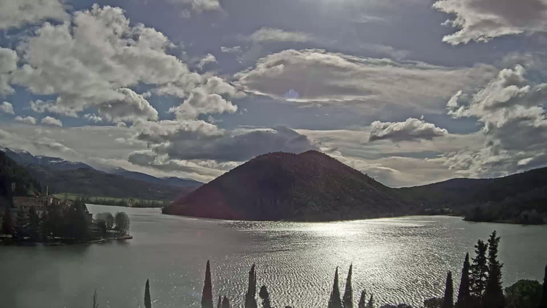 A partly cloudy day reveals a lake reflecting sunlight, with a tree-covered hill rising from its waters, backed by mountains and a small island with buildings.