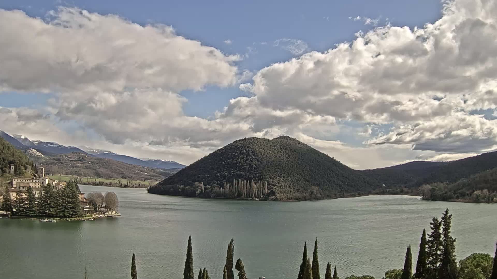A lake with a large, tree-covered hill in the middle, a village on the left shore, and snow-capped mountains in the background under a partly cloudy sky.