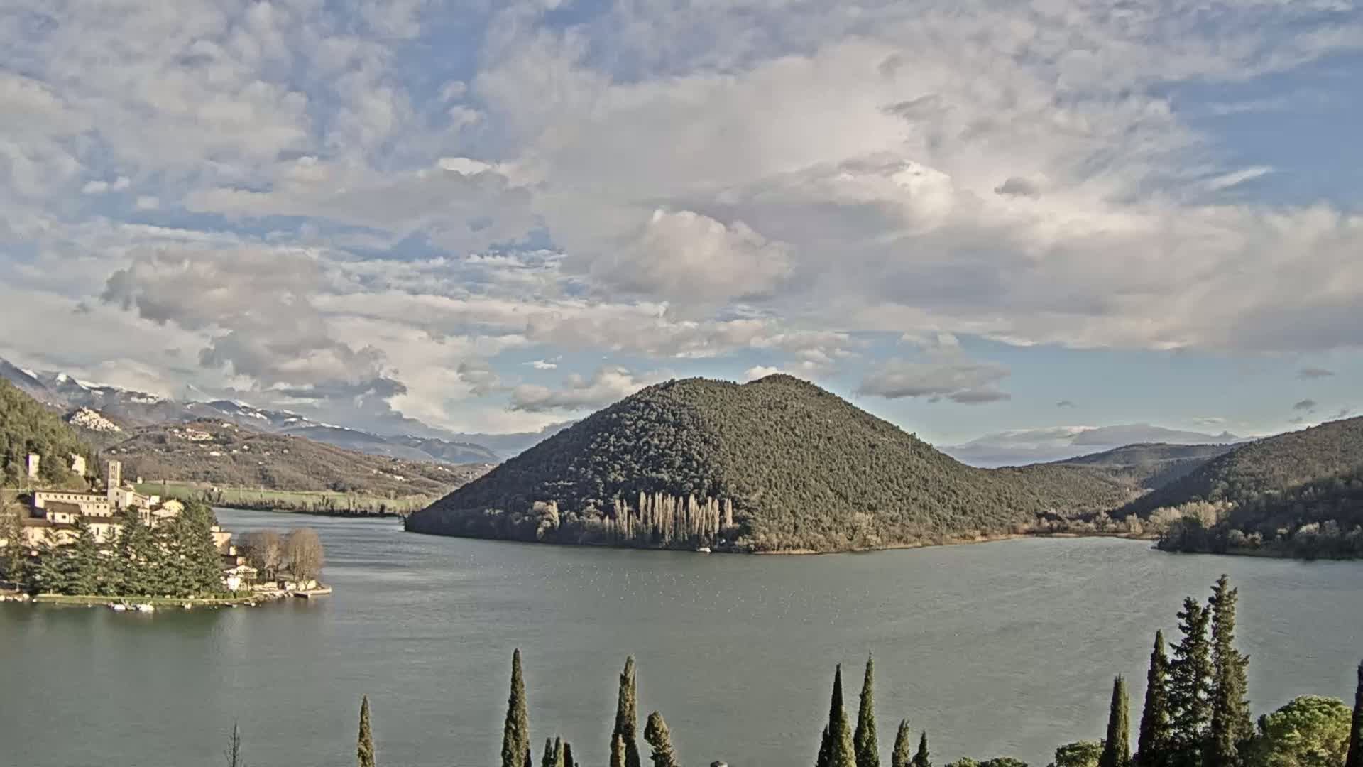 A lake with a tree-covered island and a village on its shore is seen under a partly cloudy sky, with snow-capped mountains in the distance.