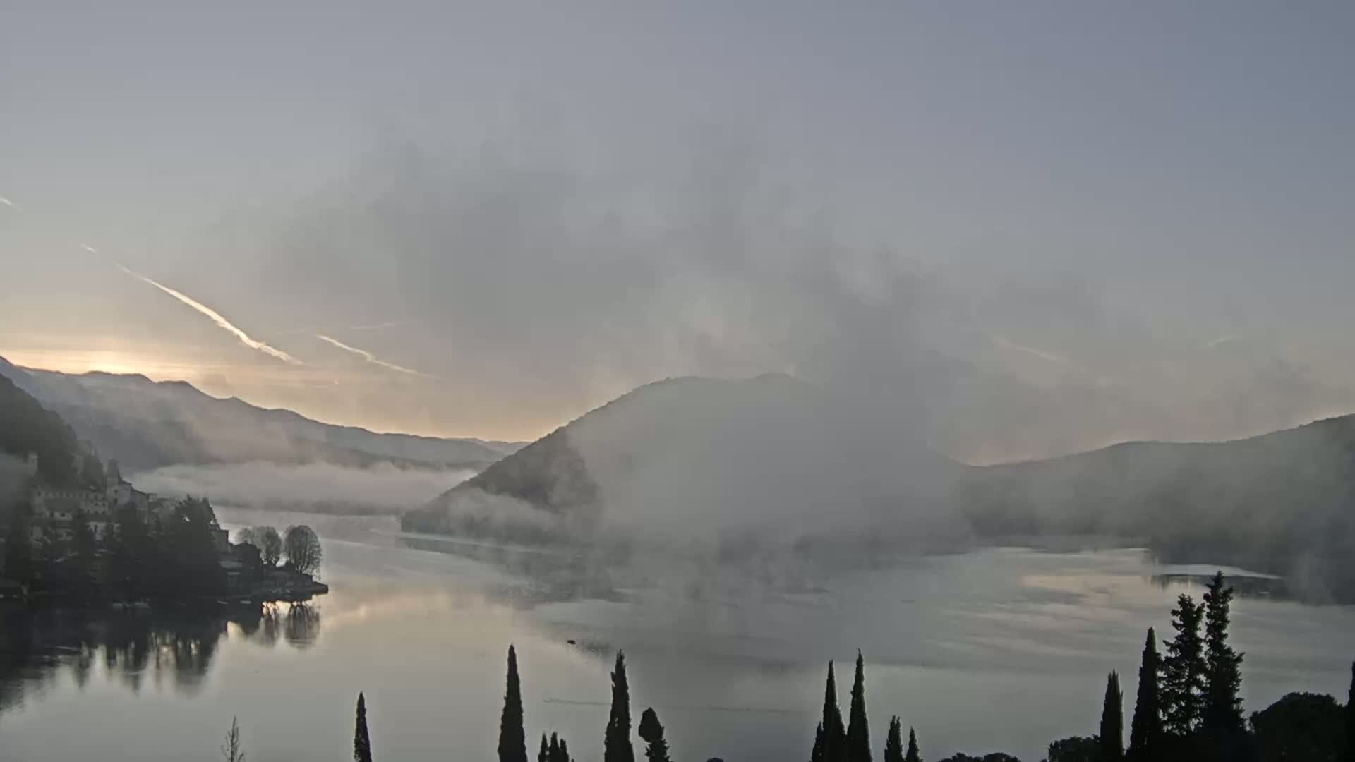 A calm lake at sunrise is shrouded in fog, with misty mountains and a small village visible in the distance.