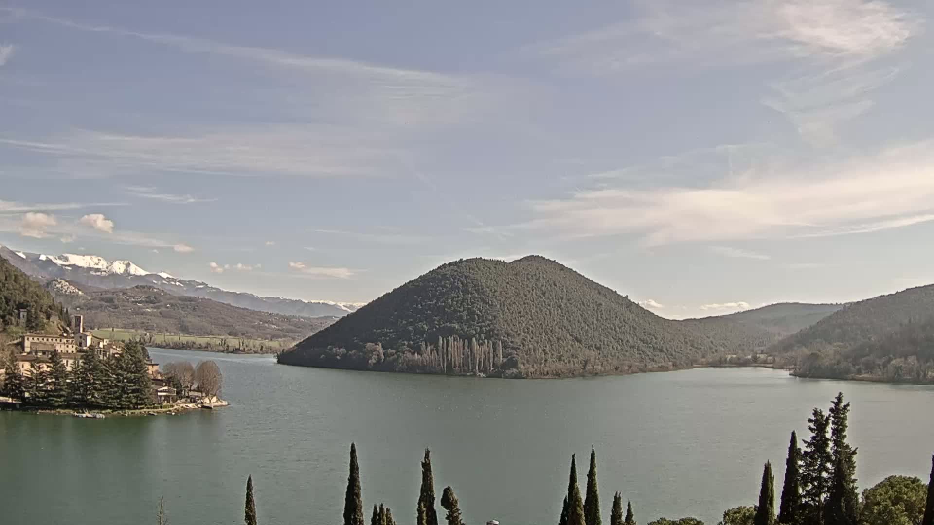 A lake surrounded by hills and mountains, some with snow-capped peaks, under a partly cloudy sky.