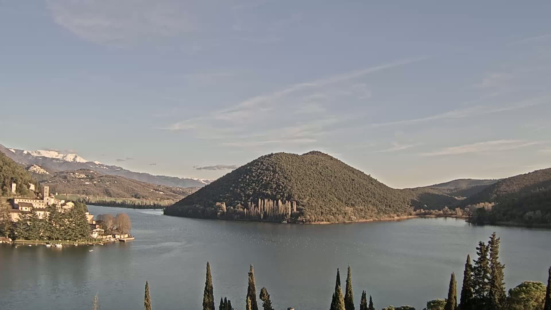 A calm lake is surrounded by hills and mountains, some of which are snow-capped under a mostly clear sky.