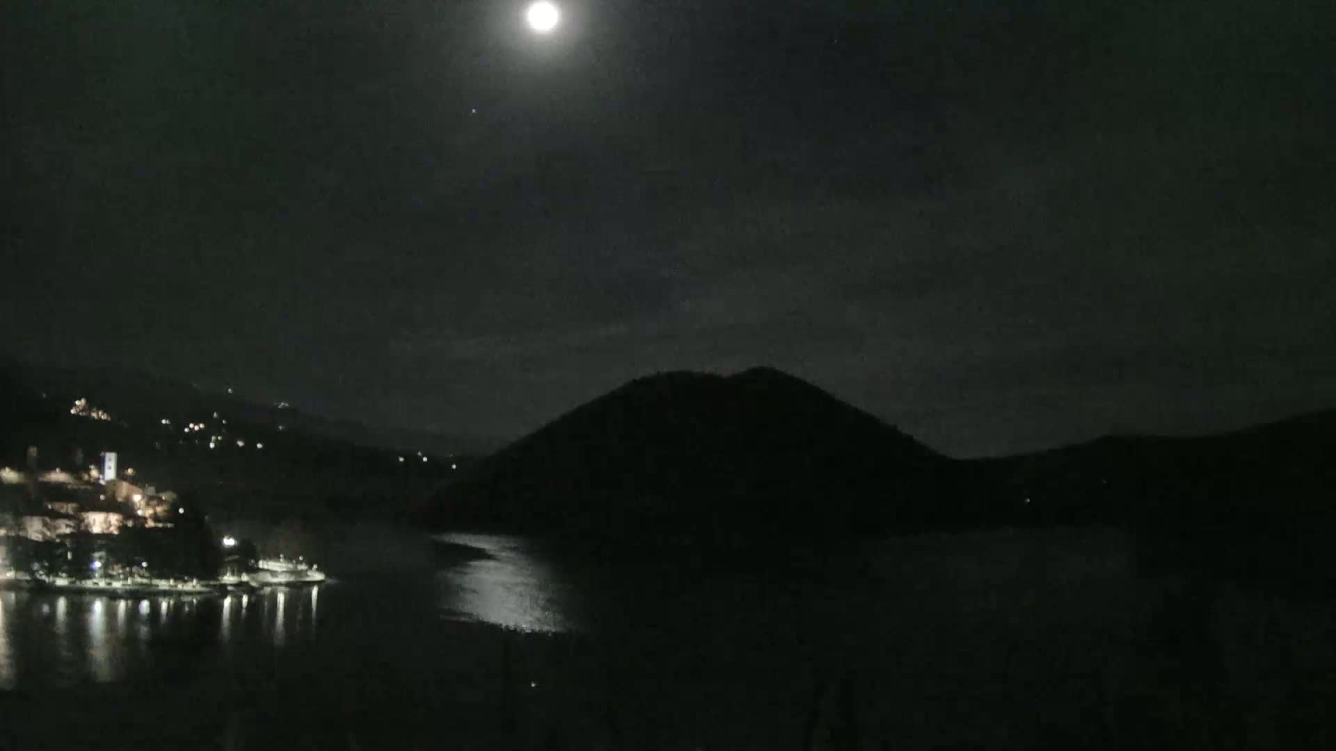 A moonlit night scene shows a body of water reflecting the light, with a dark hill silhouetted against the sky and a town's lights visible on the opposite shore.