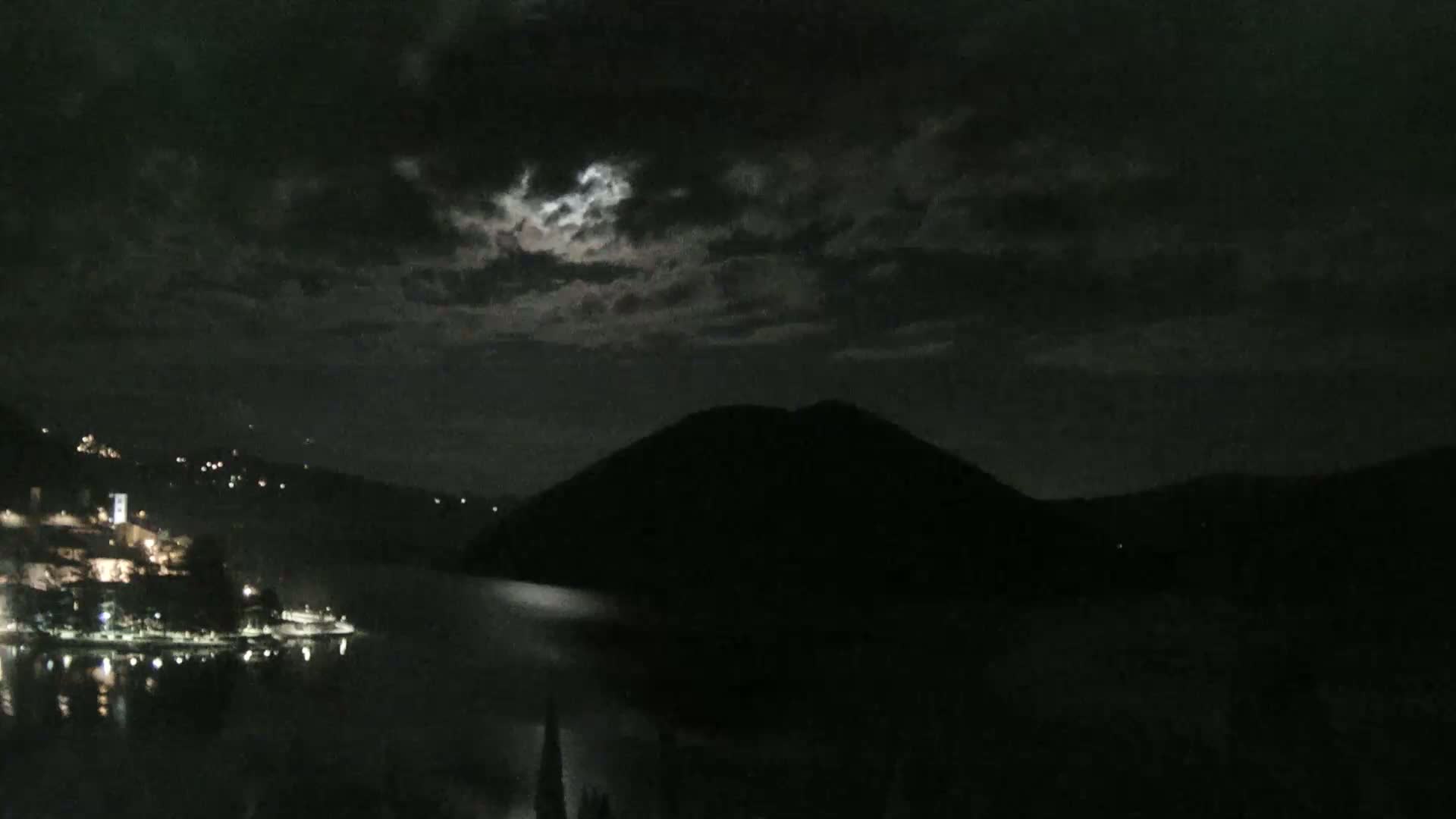 A nighttime view of a moonlit lake with a town and a dark mountain in the background under a cloudy sky.