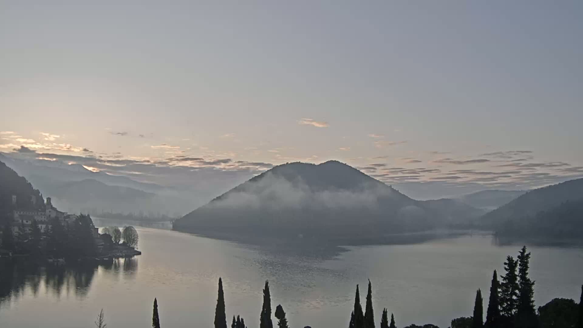 A calm lake at sunrise is surrounded by misty mountains and a small village, with a few evergreens in the foreground.