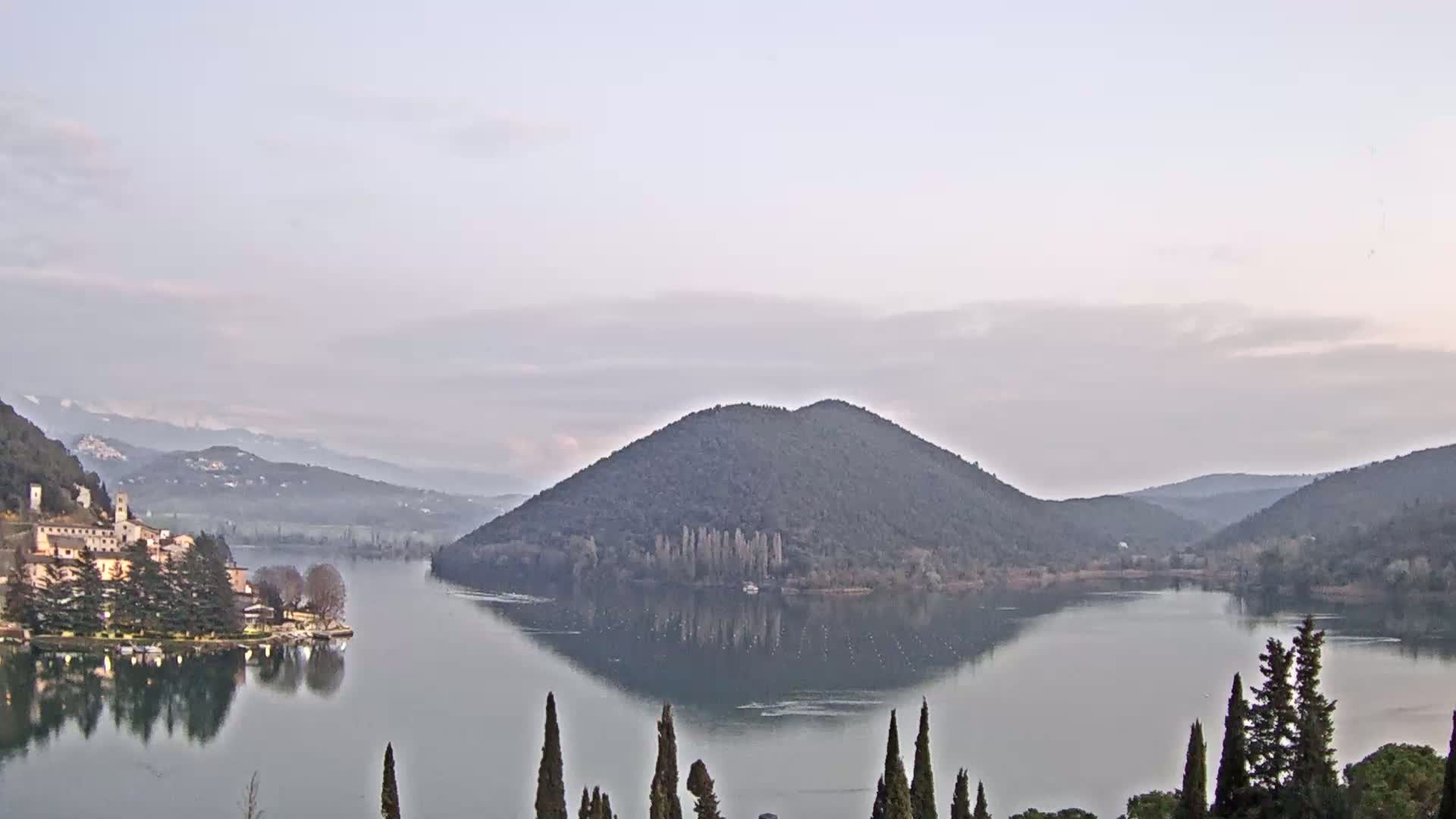 A calm lake reflects a tree-covered hill and a village nestled in the mountains under a pale sky.