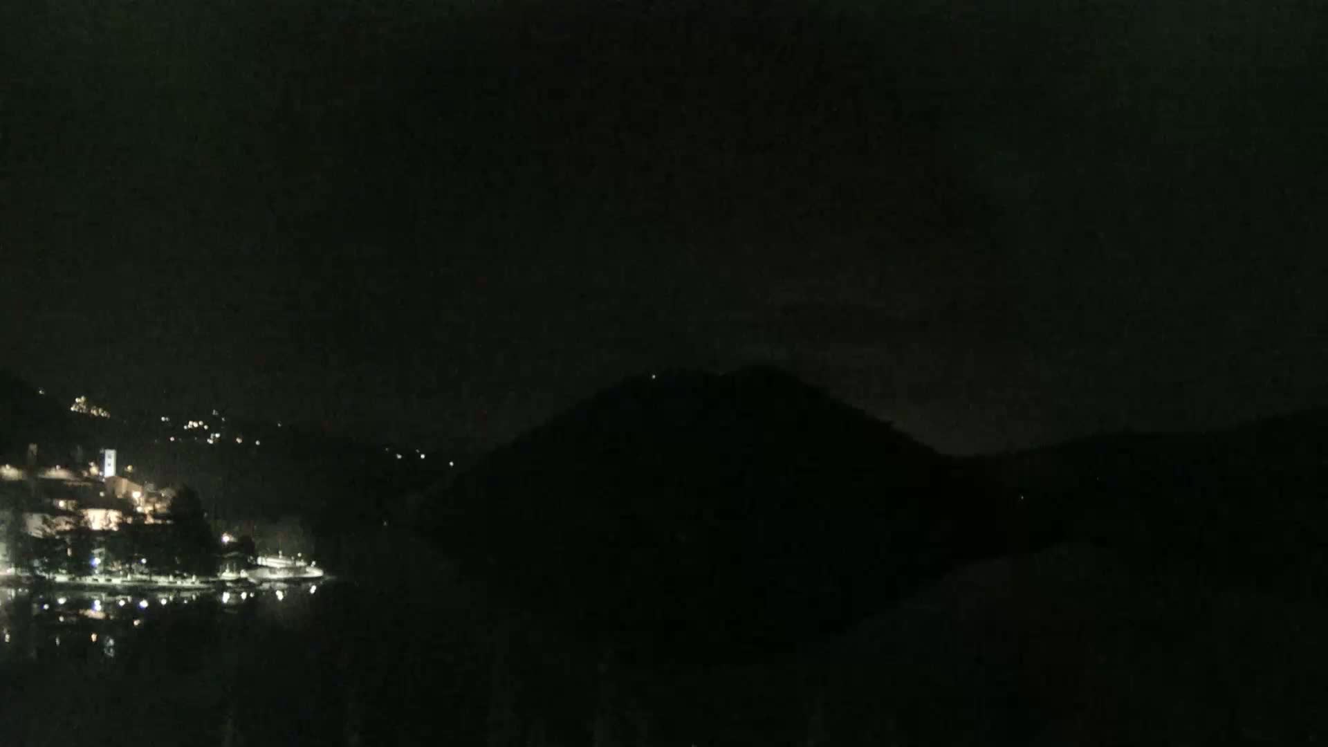 A nighttime view shows a dark, silhouetted hill overlooking a body of water with faintly lit buildings on the opposite shore under a dark sky.