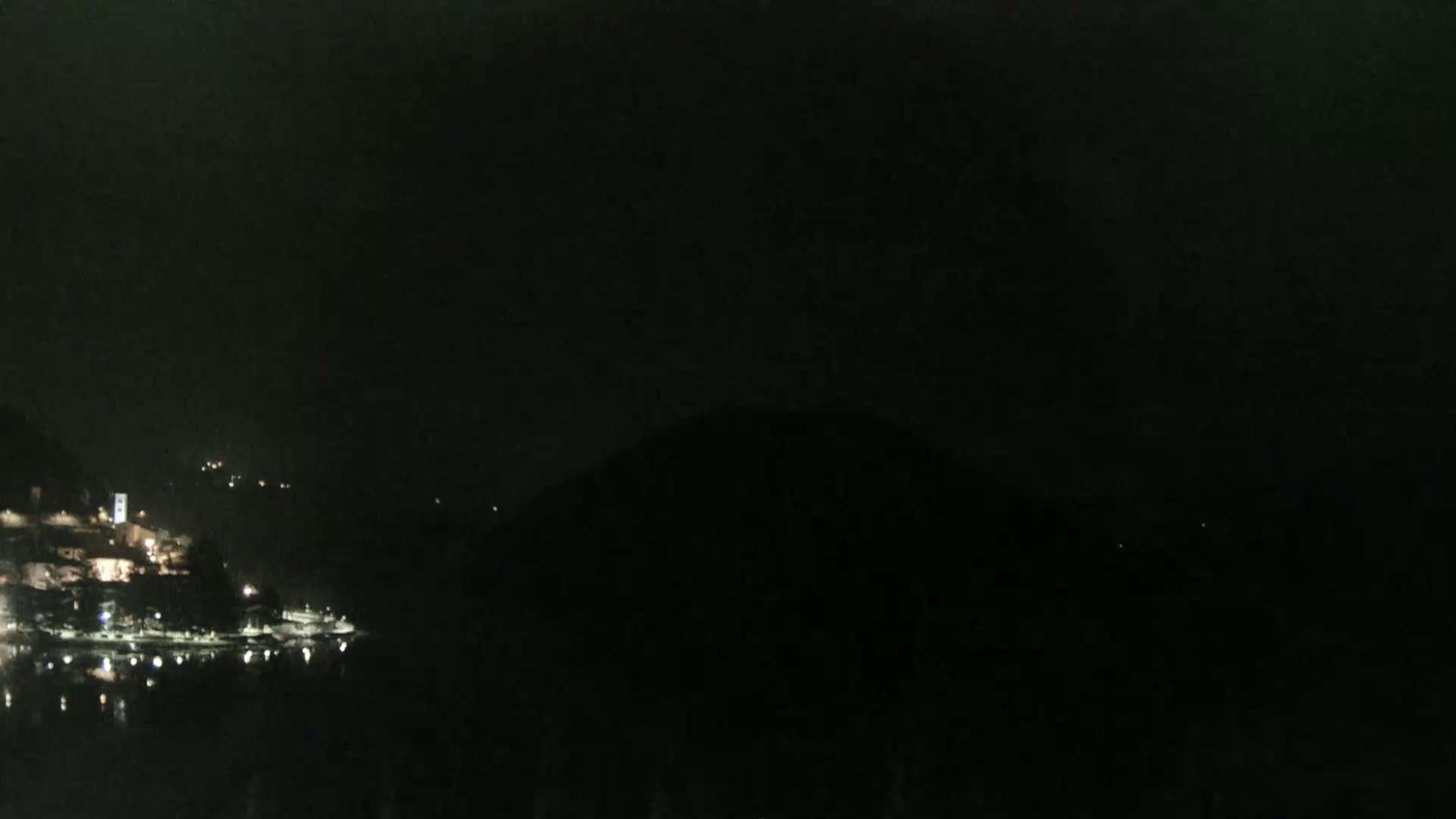 At night, a dark body of water reflects the sparse lights of a town nestled against a hill.