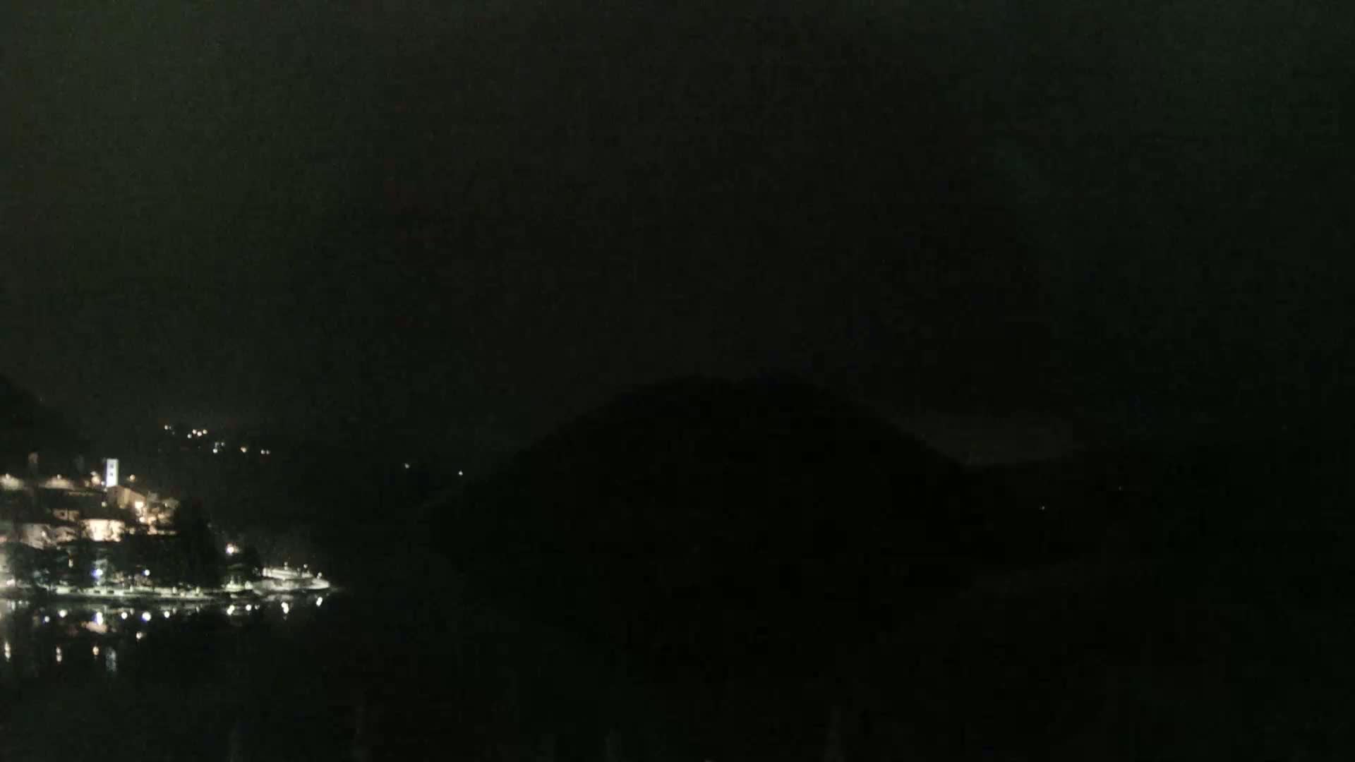 A dark nighttime scene shows a body of water reflecting a few lights from a small town nestled between dark hills.