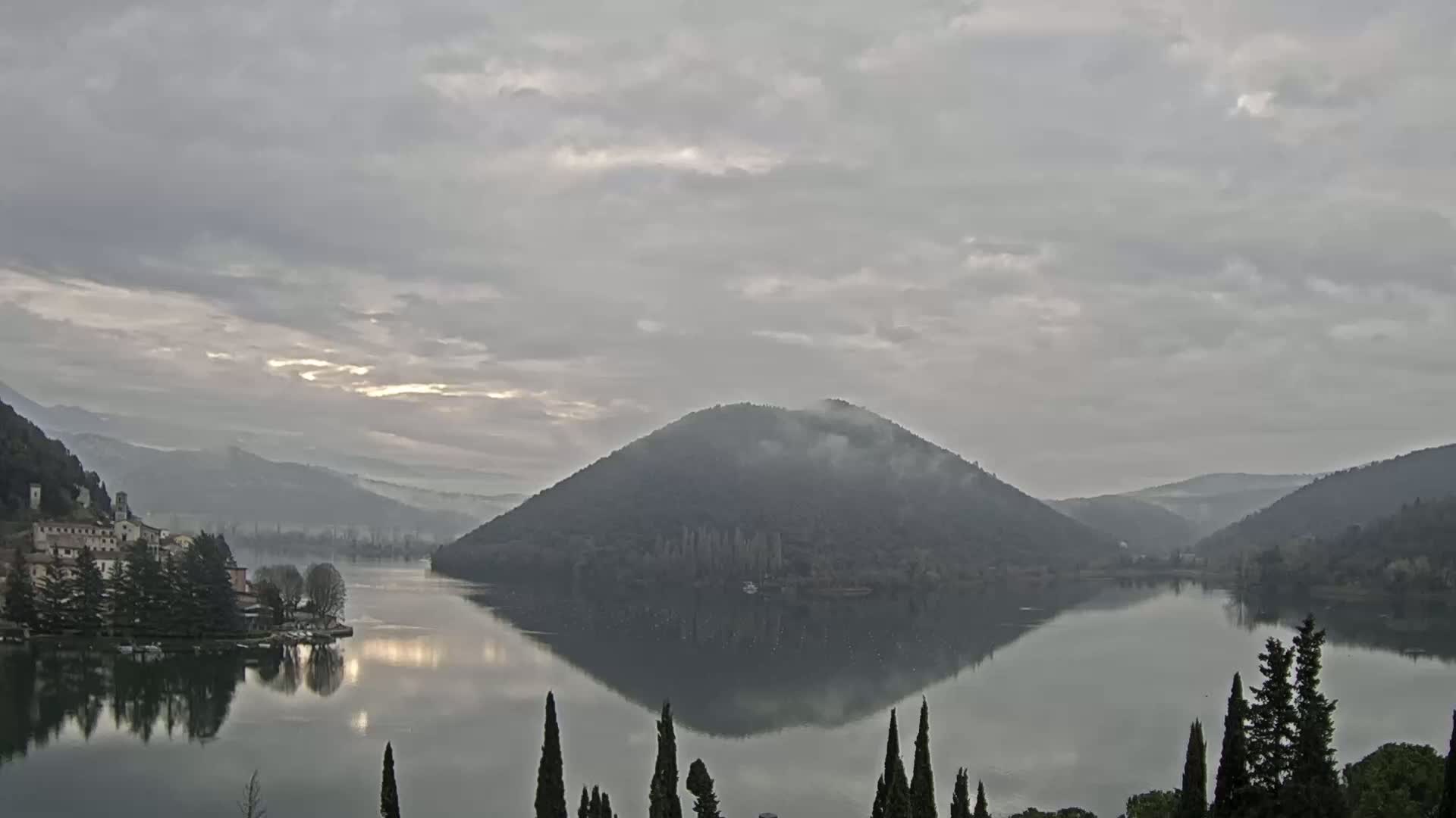 A calm lake reflects a misty, tree-covered hill and surrounding mountains under a cloudy sky.