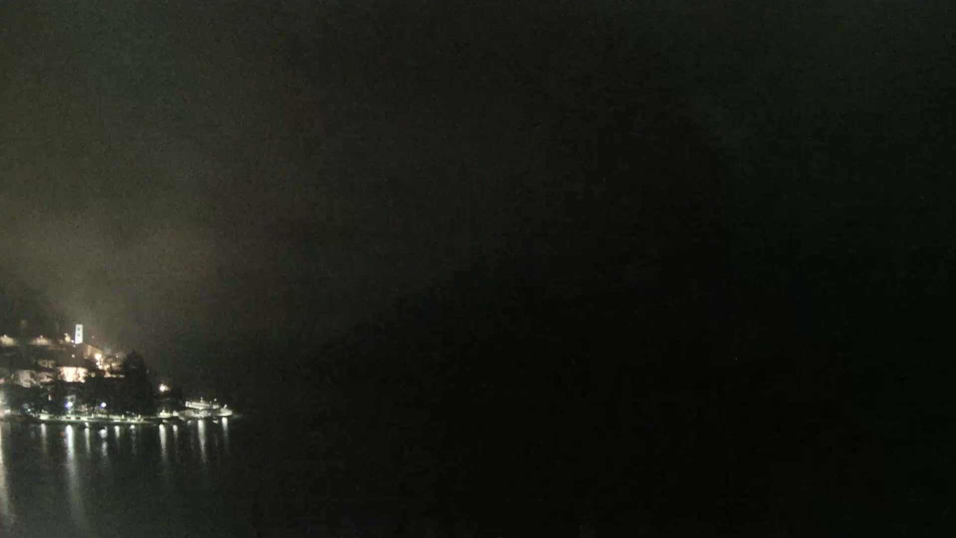 A nighttime view of a dimly lit waterfront town, partially obscured by fog or mist, under a dark sky.