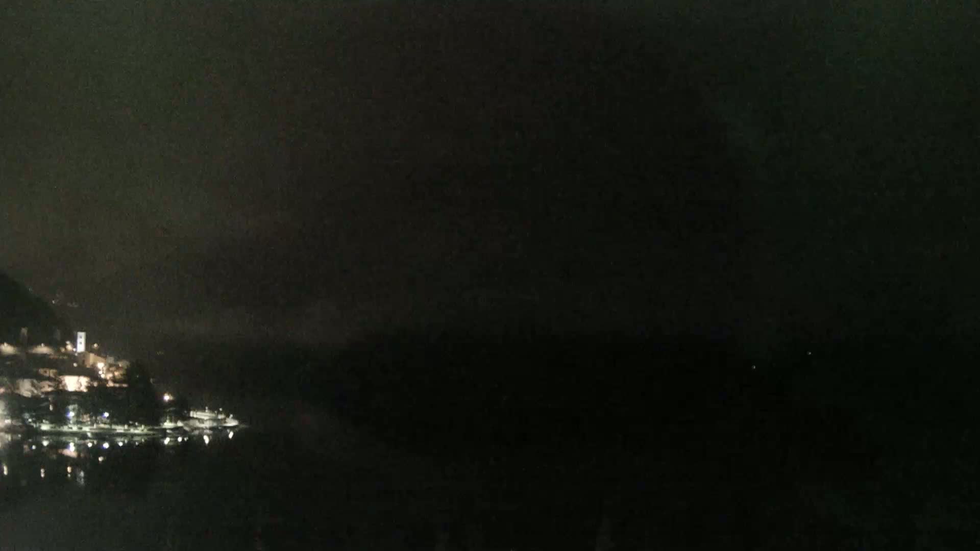 A nighttime view shows a dark body of water reflecting the faint lights of a small town nestled against a dark hillside under a cloudy, dark sky.