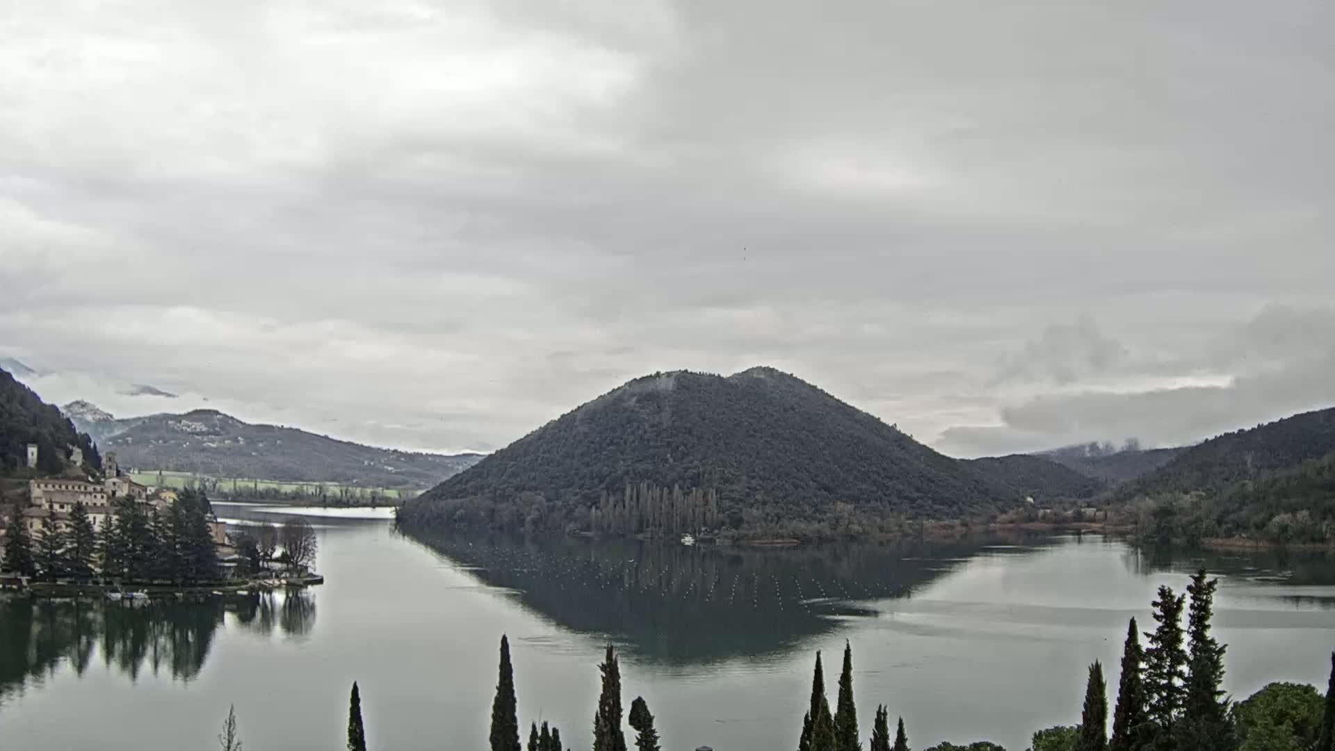 A calm lake is surrounded by dark green hills under an overcast sky, with a small town visible on the left shore and a forested island in the center.