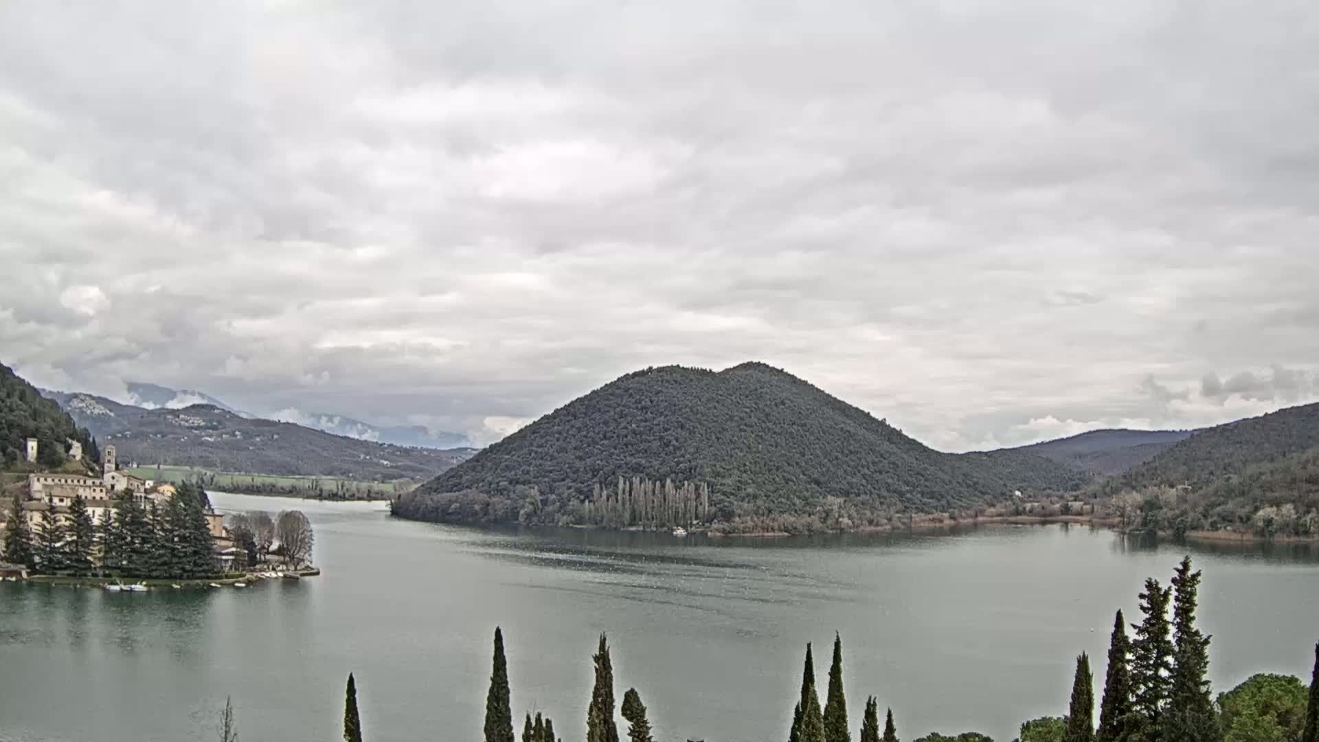 A lake is surrounded by hills and mountains under an overcast sky, with a small village visible on the left shore.