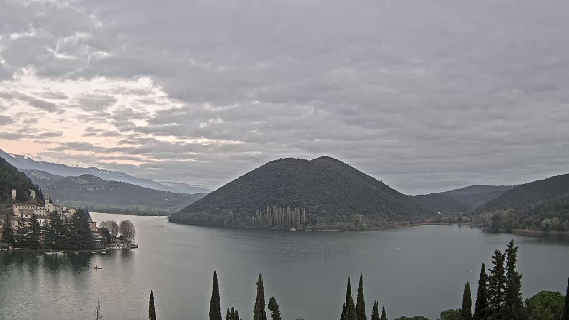 A lake is surrounded by hills and mountains under a cloudy sky, with a small town visible on one shore.