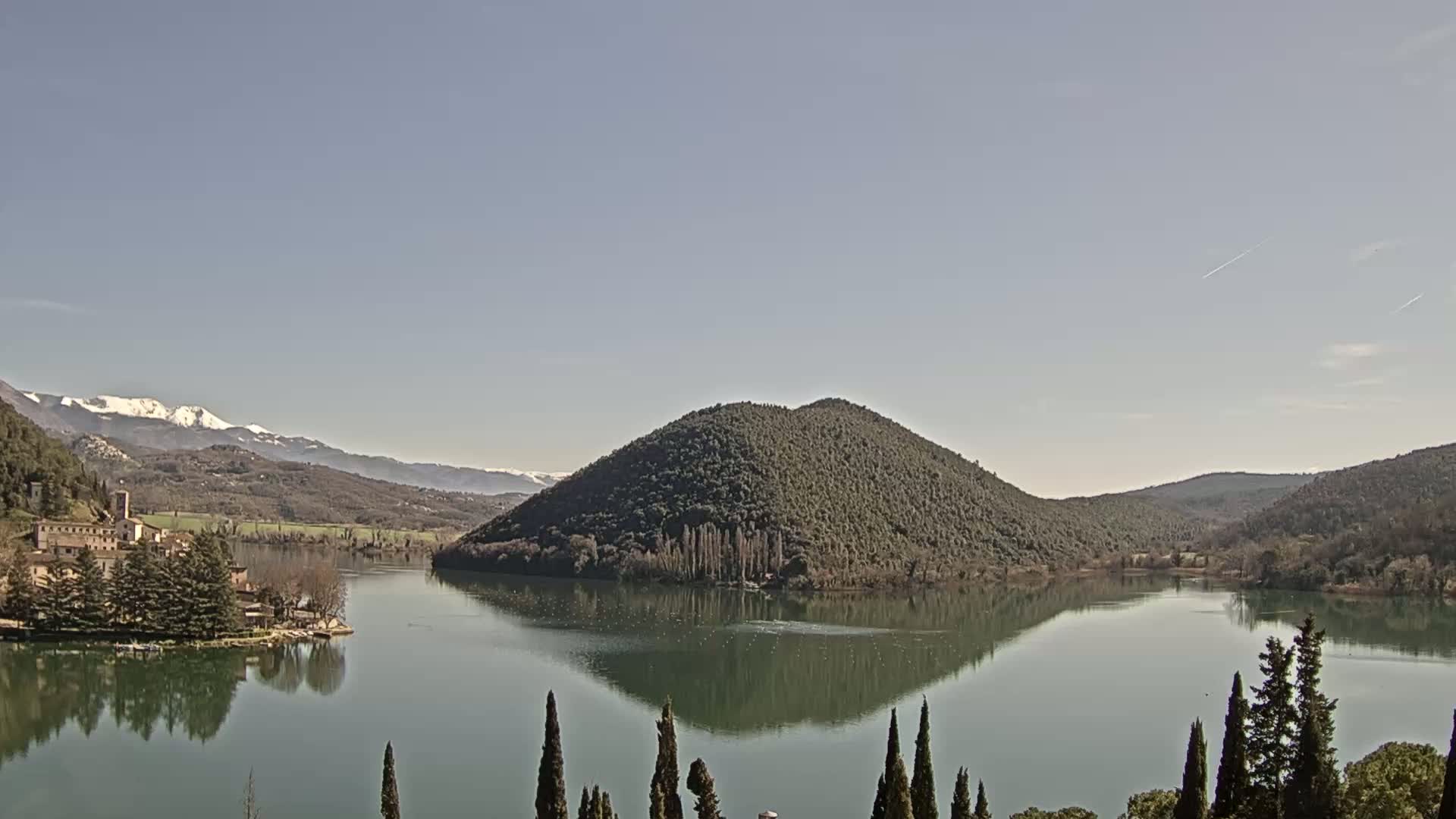 A calm lake reflects a tree-covered hill and snow-capped mountains under a clear sky.