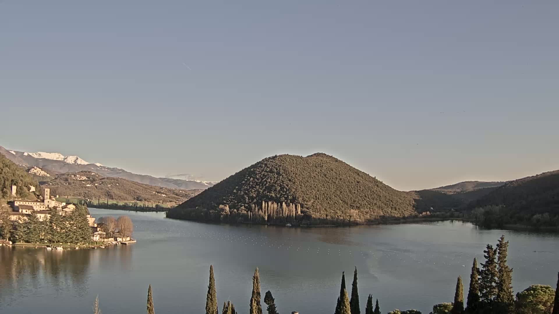 A calm lake is surrounded by hills and mountains, some of which are snow-capped under a clear sky.