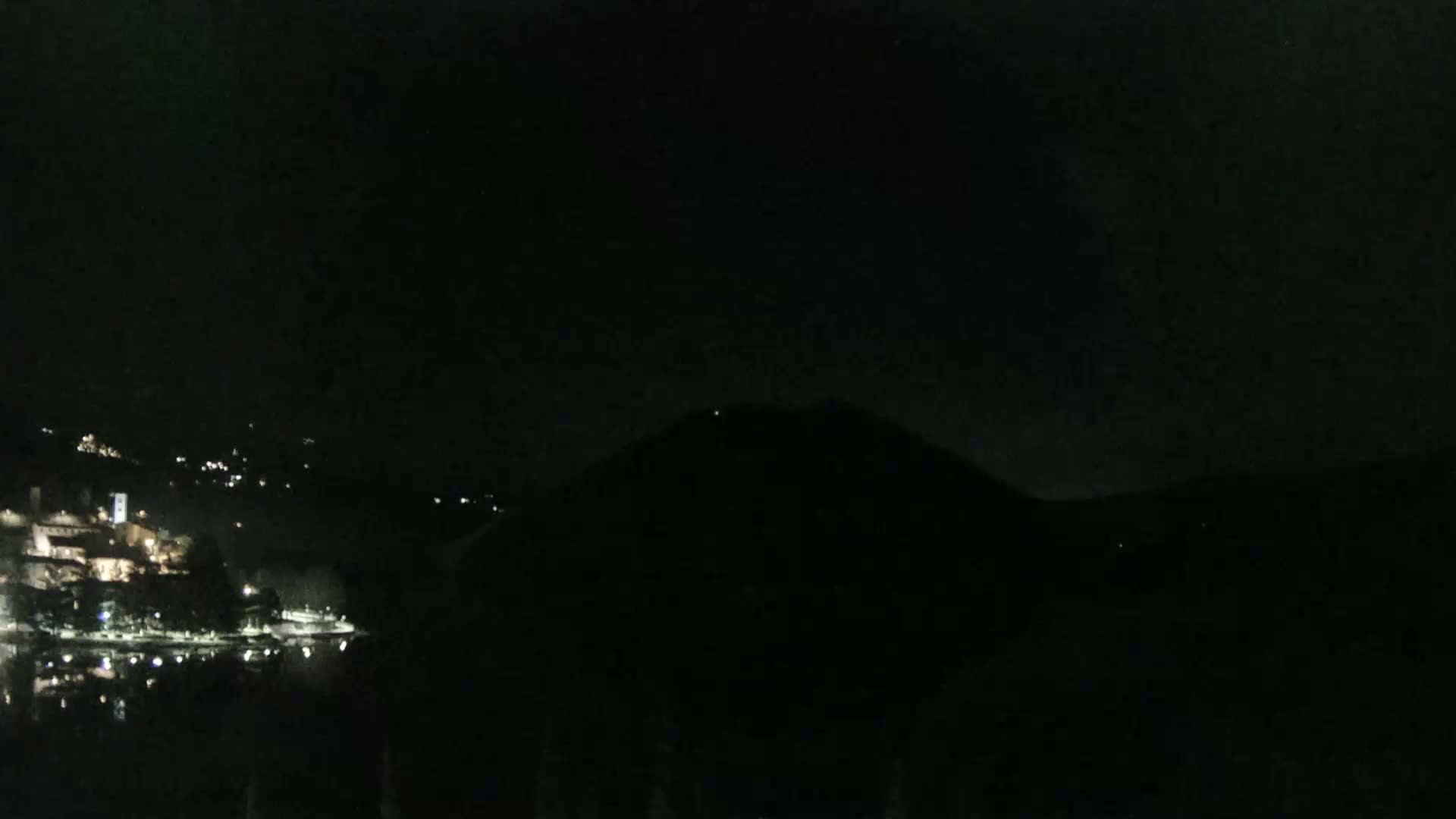 A dark, nighttime view shows a body of water reflecting the lights of a town nestled in the hills across the water, with a large, dark hill or mountain in the foreground.