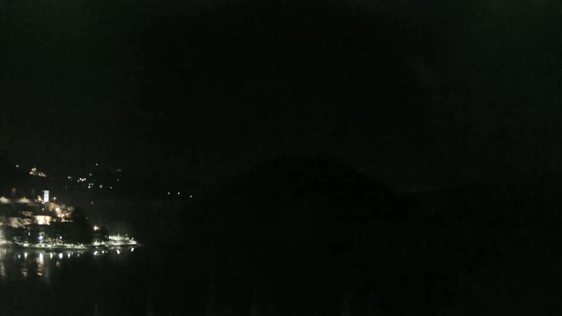 A dark, nighttime scene shows a body of water reflecting the lights of a town on the opposite shore, under a clear, dark sky.
