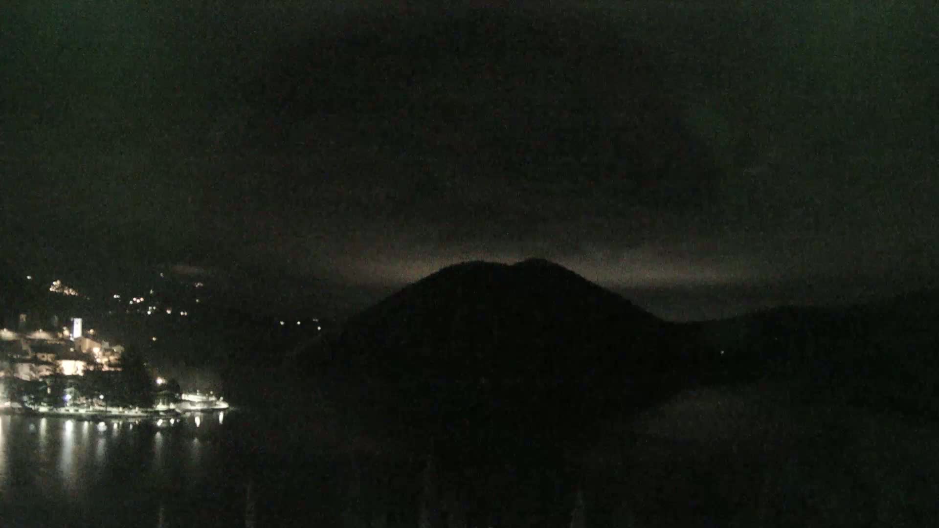 A dark, nighttime scene shows a body of water reflecting the lights of a town nestled at the base of a silhouetted mountain under a mostly cloudy sky.