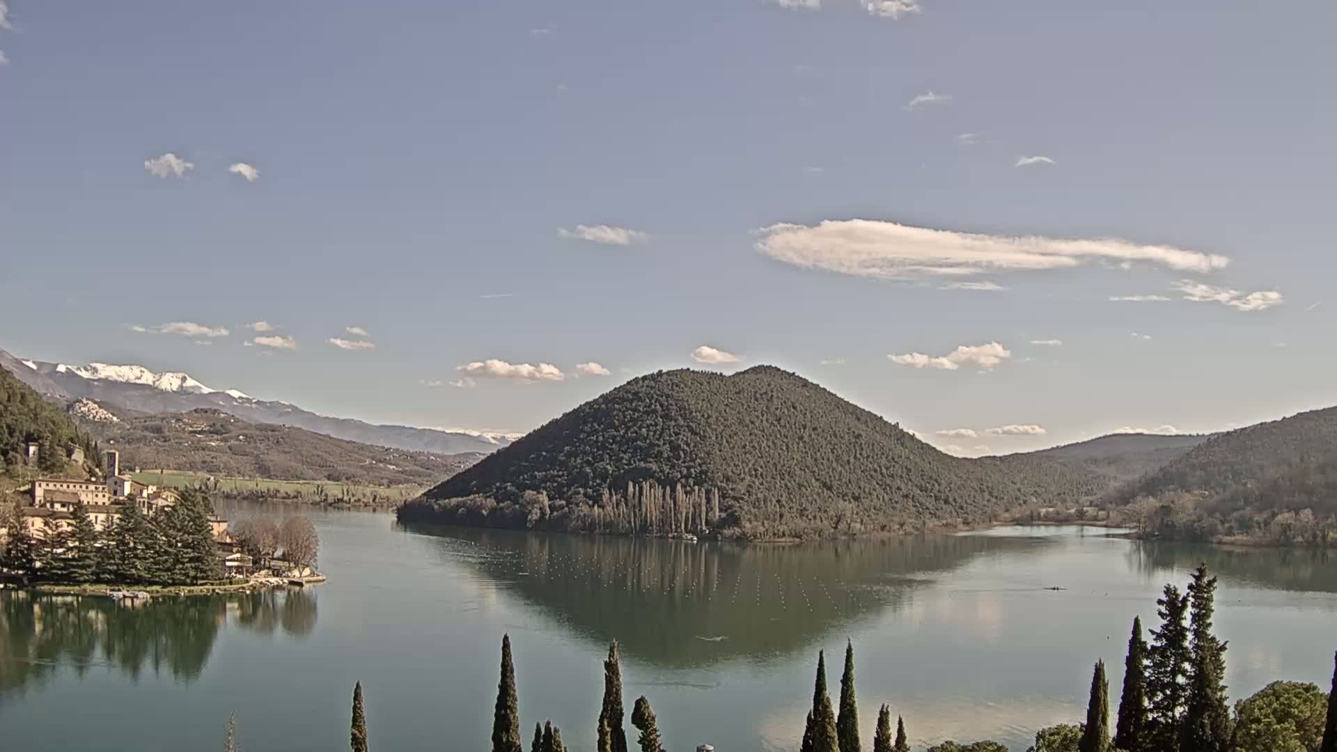 A calm lake reflects a tree-covered hill and snow-capped mountains under a mostly clear sky with a few scattered clouds.