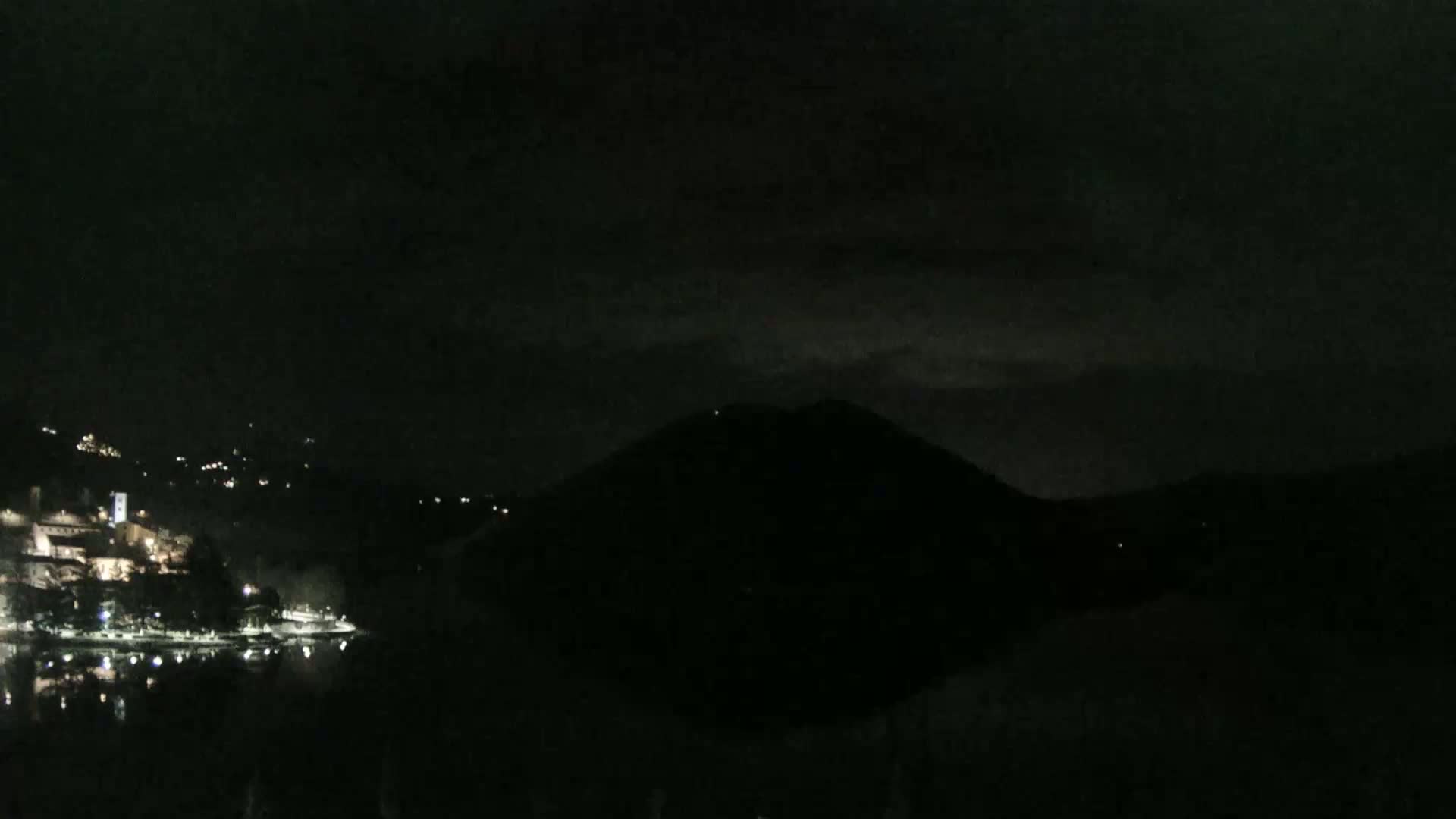 A dark, nighttime scene shows a body of water reflecting a dimly lit lakeside town, with a large, dark hill or mountain rising from the water's edge under a mostly cloudy sky.