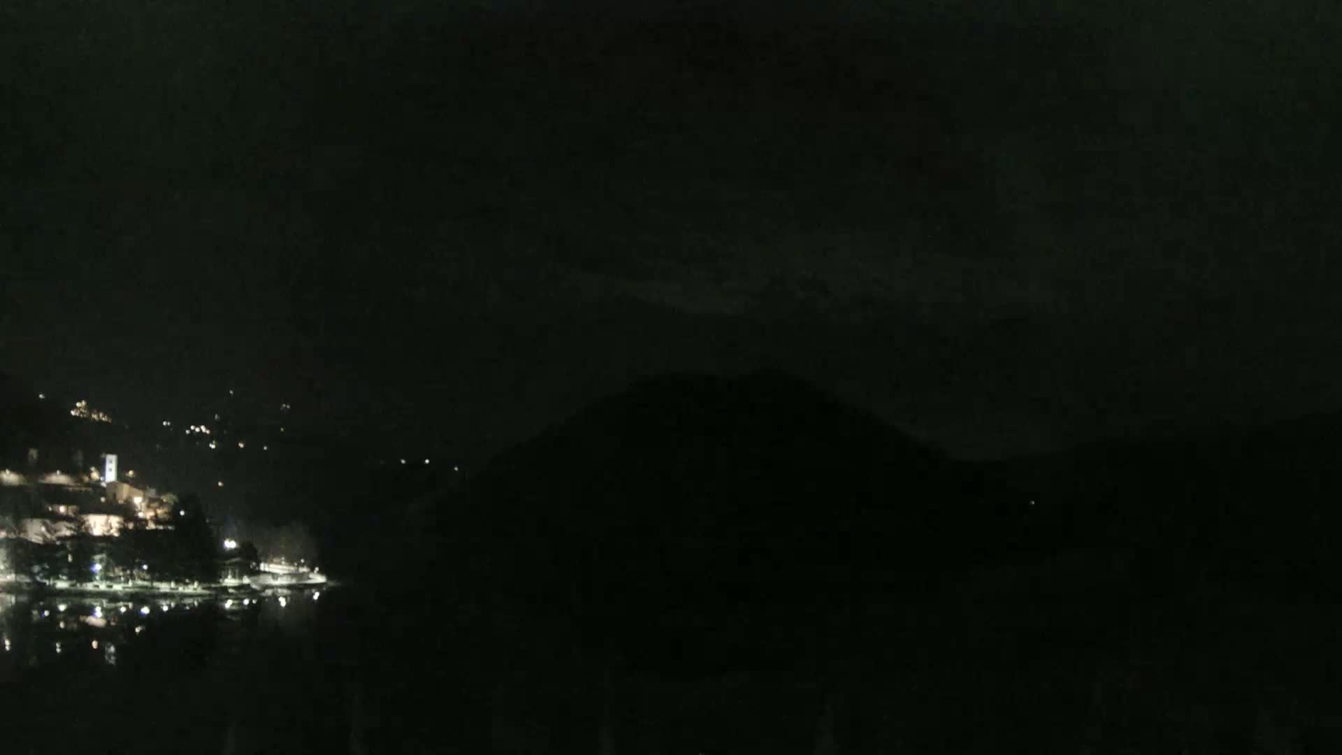 A nighttime view of a lakeshore town with sparsely lit buildings, partially obscured by darkness and a hill, under a dark, cloudy sky.