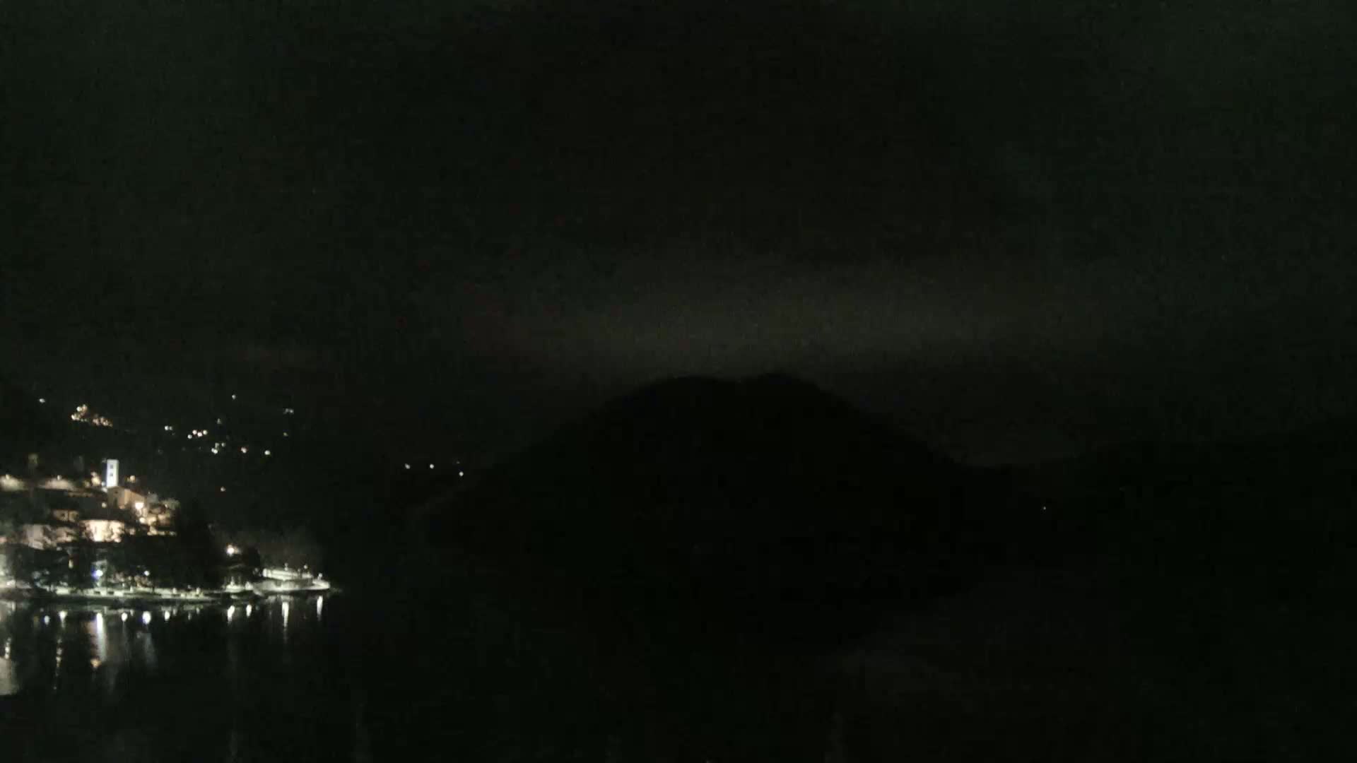 A nighttime view shows a dark, hilly landscape across a body of water reflecting scattered lights from a town on the opposite shore under a cloudy sky.